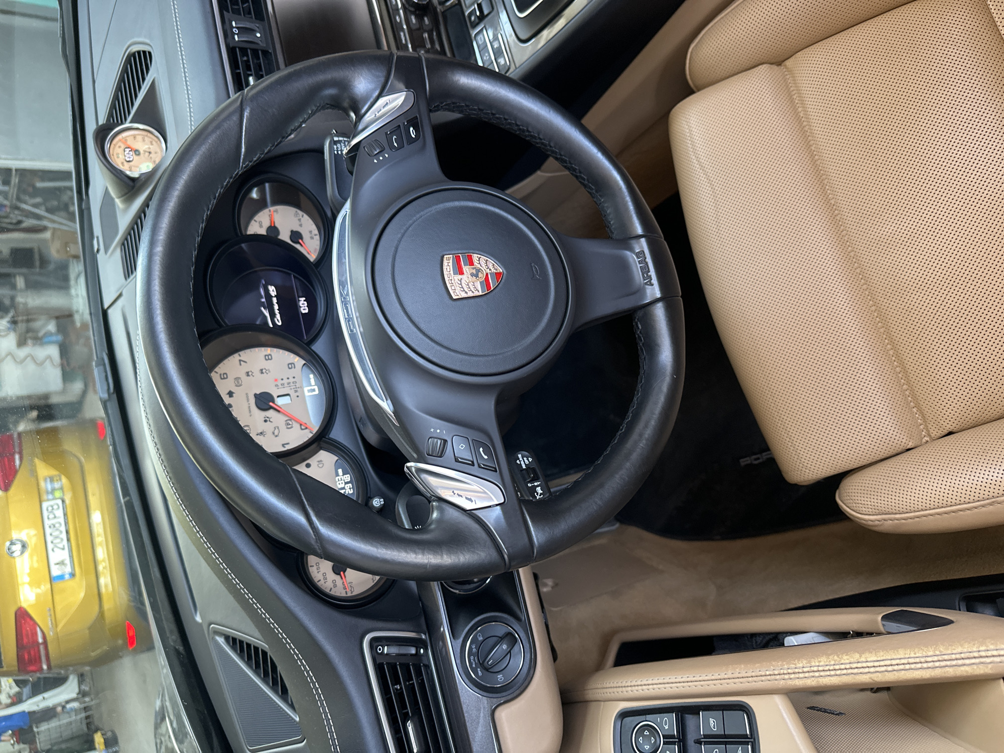 This media file is a close-up, interior shot of a car, likely a Porsche, with a focus on the dashboard and steering wheel. The dominant subjects are the car's interior elements: a black leather steering wheel with a Porsche emblem, a dashboard with multiple gauges and displays, and a tan leather seat. The car's interior appears to be in good condition, with clean lines and stitching visible on the leather.
In the background, a bright yellow car is visible through the car's windshield or side window, suggesting the vehicles are either in a dealership, a garage, or a similar setting. The yellow car has a license plate that reads "A 2008 PB".
There is no specific activity or event taking place; the image seems to be a still shot of the car's interior. Notable details include the various gauges on the dashboard, one of which appears to display "Carrera 4S". The text "AIRBAG" is clearly visible on the steering wheel. The lighting suggests it could be daytime, but the exact time of day and weather are not discernible. The overall impression is one of a well-maintained, luxury vehicle interior.