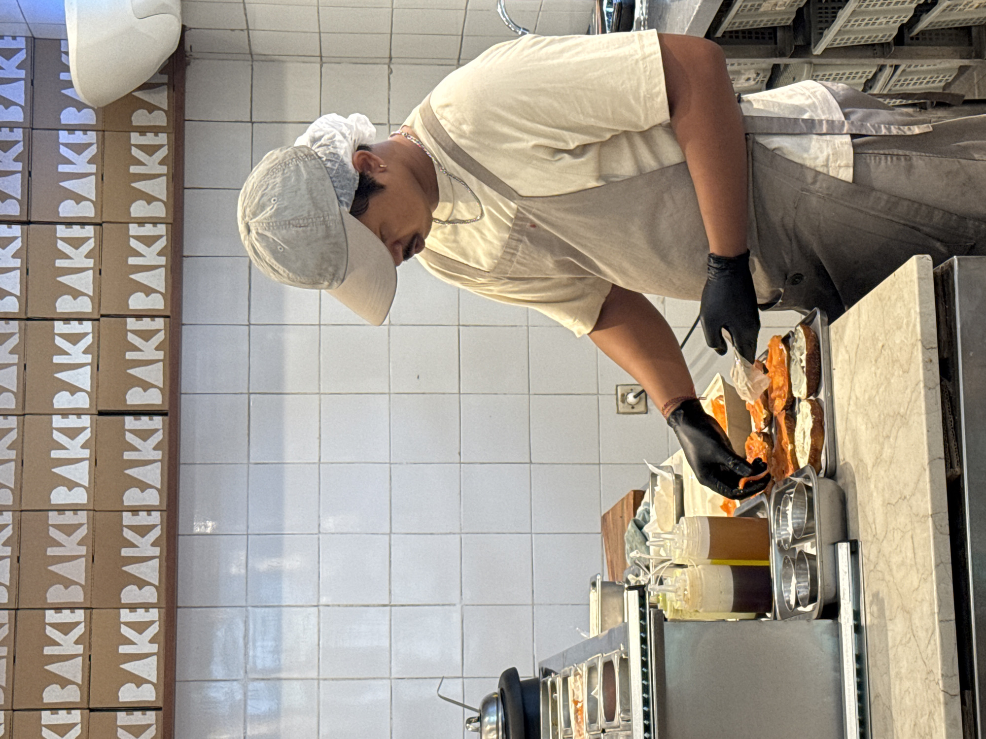 Inside a commercial kitchen, an individual wearing a light beige short-sleeved shirt, an apron, a hair net, and a light gray hat is preparing food. They are using black gloves while arranging food items on a tray. To their left, boxes with the word "BAKE" printed repeatedly in white text are stacked vertically. The kitchen features white tiled walls and includes stainless steel equipment and ingredient containers. The image was taken in North Kuta, Indonesia.
