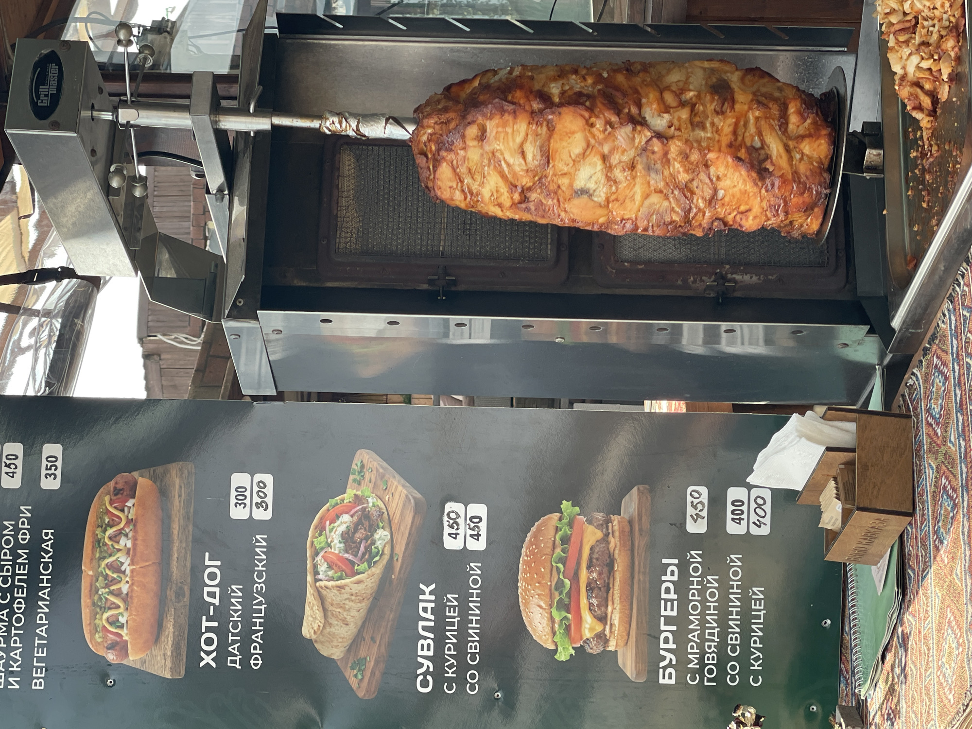 This image shows an outdoor food stall in Sochi, Russia. The primary subject is a vertical rotisserie with a large piece of seasoned meat slowly cooking. In the foreground, a menu board displays various food items with their prices, including shawarma, hot dogs, souvlaki, and burgers. The menu is written in Russian. The stall appears to be busy, with food being prepared and served. The overall scene is one of a casual, bustling food establishment.