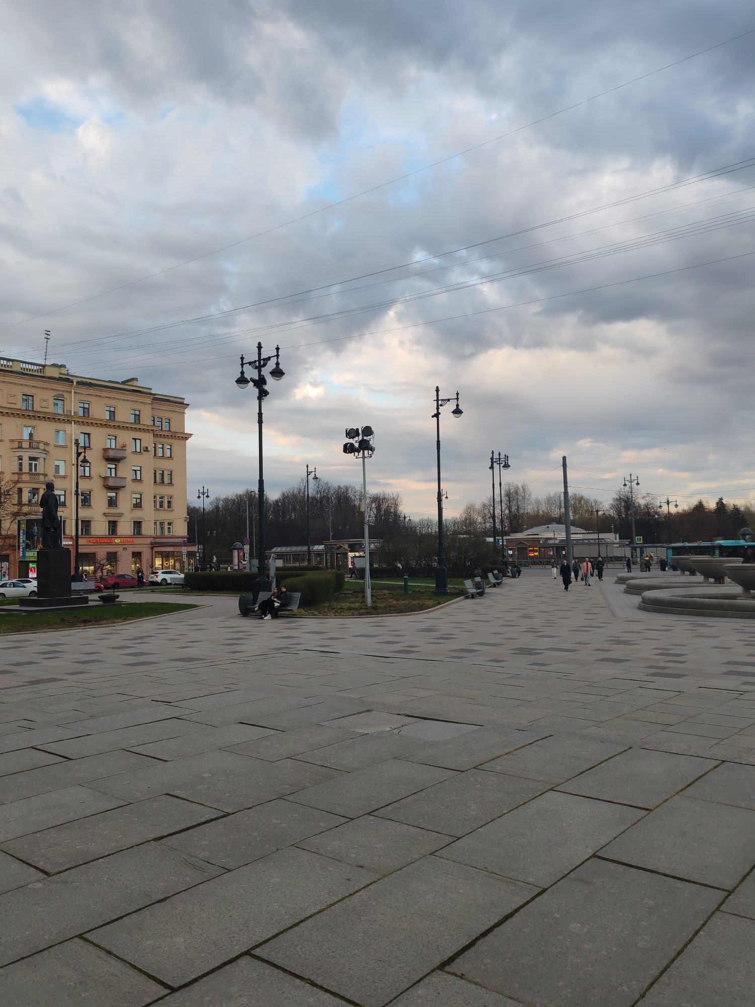 This image captures a wide urban square or plaza, likely in Saint Petersburg, Russia, under a cloudy, overcast sky with hints of blue. The time of day appears to be late afternoon or early evening, characterized by subdued light.
In the foreground, the ground is covered with a large expanse of grey and dark grey paving stones laid in rectangular and square patterns. To the left, a prominent multi-story building with a light beige facade, numerous windows, and balconies stands alongside a street where a few cars are visible, including a red one. A dark statue on a pedestal is positioned in front of this building. On the ground floor of the building, a shop sign in Cyrillic is discernible, reading "Счастливый взгляд" (Schastlivyy vzglyad), which translates to "Happy Look" or "Happy Glance," indicating an optical store.
Several tall, black ornate streetlights with multiple lamps are spaced throughout the scene. One such streetlight stands near the center, beneath which a couple is seated on a bench, next to a patch of grass. Further into the midground and to the right, more individuals are walking across the paved area. The landscape features numerous bare trees, suggesting a season of late autumn, winter, or early spring. To the far right, a large, multi-tiered concrete fountain structure is visible, appearing to be dry. In the distance, other low-rise buildings and a blue public transport vehicle (possibly a bus) can be seen. Numerous overhead wires stretch across the cloudy sky. The scene depicts a calm, ordinary urban setting with daily activities.