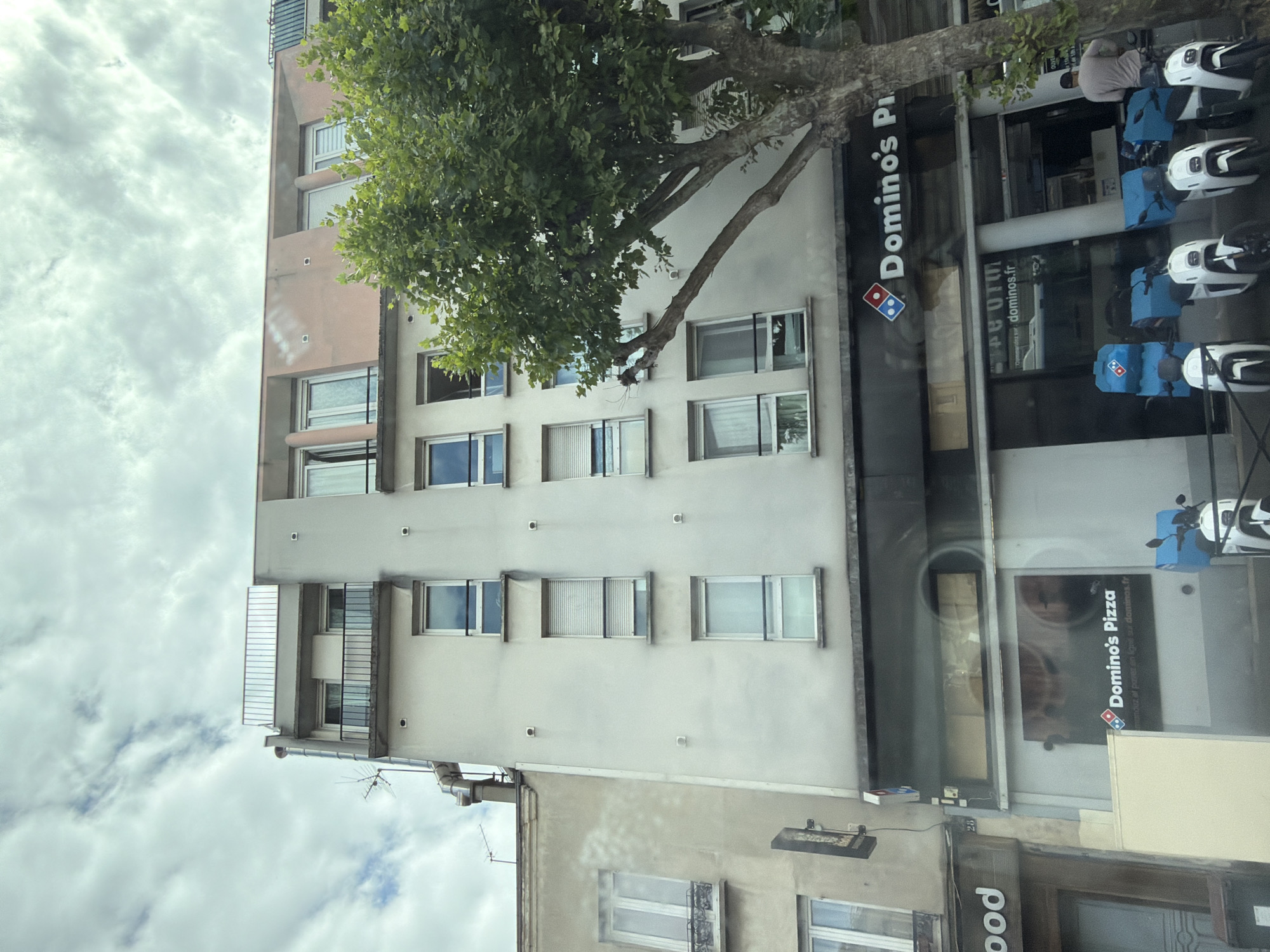 This image captures a street view of a building with a Domino's Pizza restaurant on the ground floor in Alfortville, France. The building is light-colored and has multiple stories with numerous windows. A large tree with green foliage is prominently situated to the right of the building, partially obscuring it. The Domino's Pizza storefront features its distinctive logo and branding, with a slogan that appears to be in French. In front of the restaurant, several white electric scooters are parked, some equipped with blue delivery boxes bearing the Domino's logo. A person in a light-colored shirt and dark pants is seen walking away from the scooters towards the right side of the frame. The sky is partly cloudy.