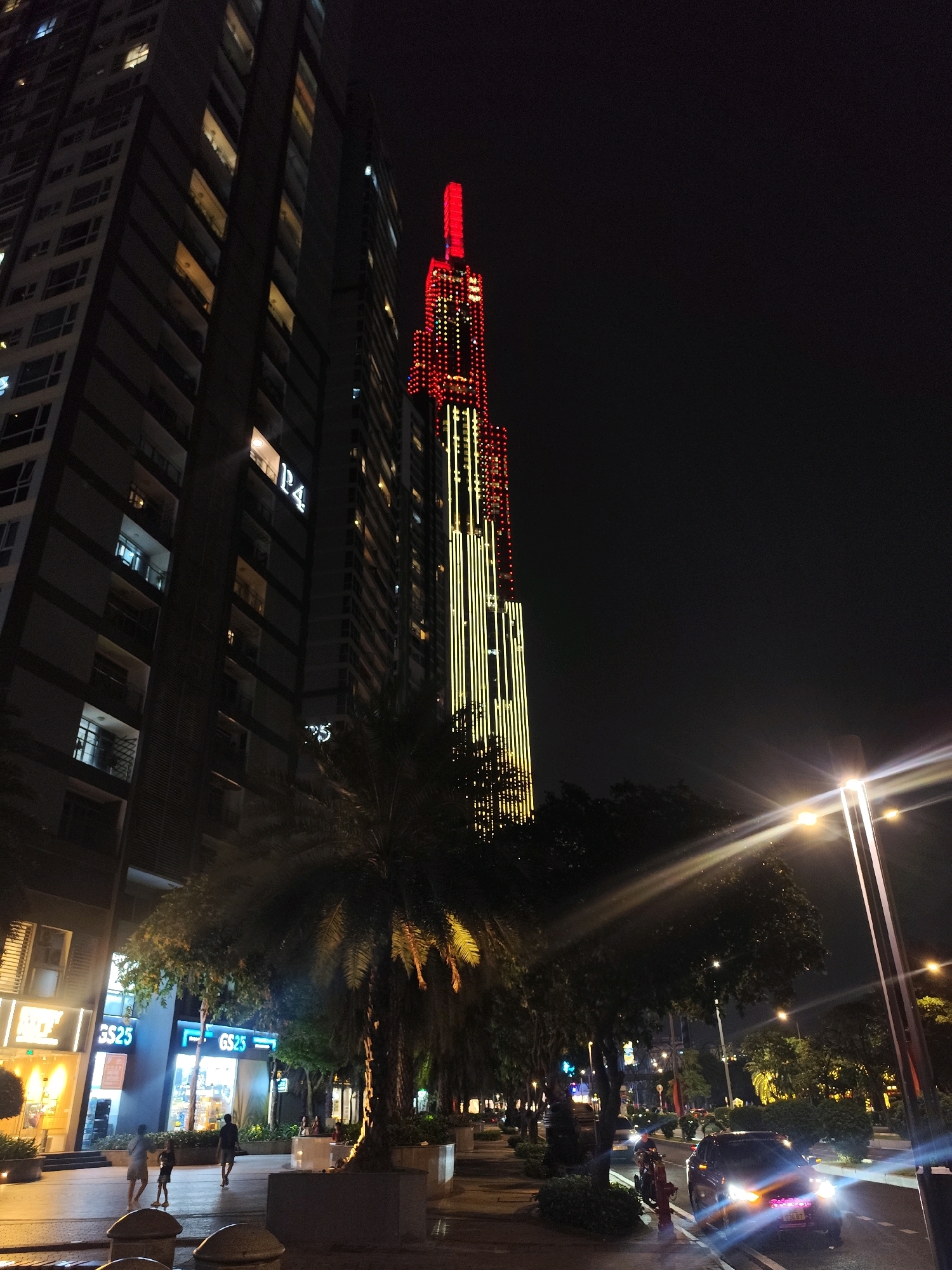This is a night-time, eye-level, outdoor, full-shot photograph of Landmark 81, a skyscraper in Ho Chi Minh City, Vietnam. The tower is illuminated with red and yellow lights, creating a vibrant spectacle against the dark sky. To the left of the tower, another tall building with illuminated windows is visible. In the foreground, a tree-lined sidewalk runs alongside a street where cars and motorcycles are driving. Streetlights cast a warm glow on the scene, and there are several people walking on the sidewalk. One of the people appears to be a child. The ground is wet, indicating recent rain, and a reflection of the lights can be seen on the pavement. A GS25 convenience store is visible on the left side of the image.