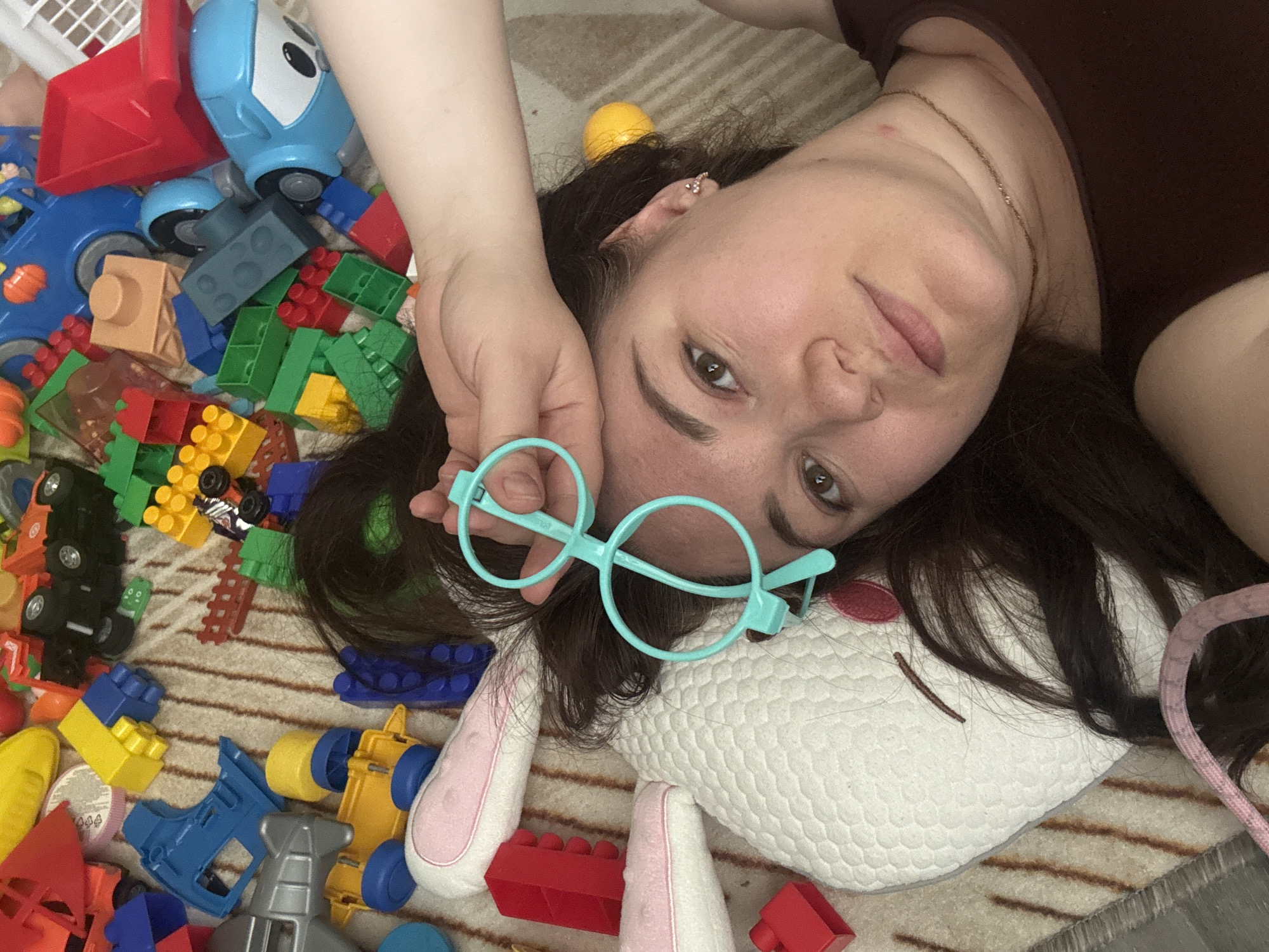 A woman is lying on a striped rug amidst various toys. The toys include building blocks, toy vehicles, and a stuffed animal resembling a rabbit. The woman is holding a pair of blue round-framed glasses in front of her face. The scene appears to be indoors. There is no evidence to confirm the location in the city of Chaikovsky, Russia.