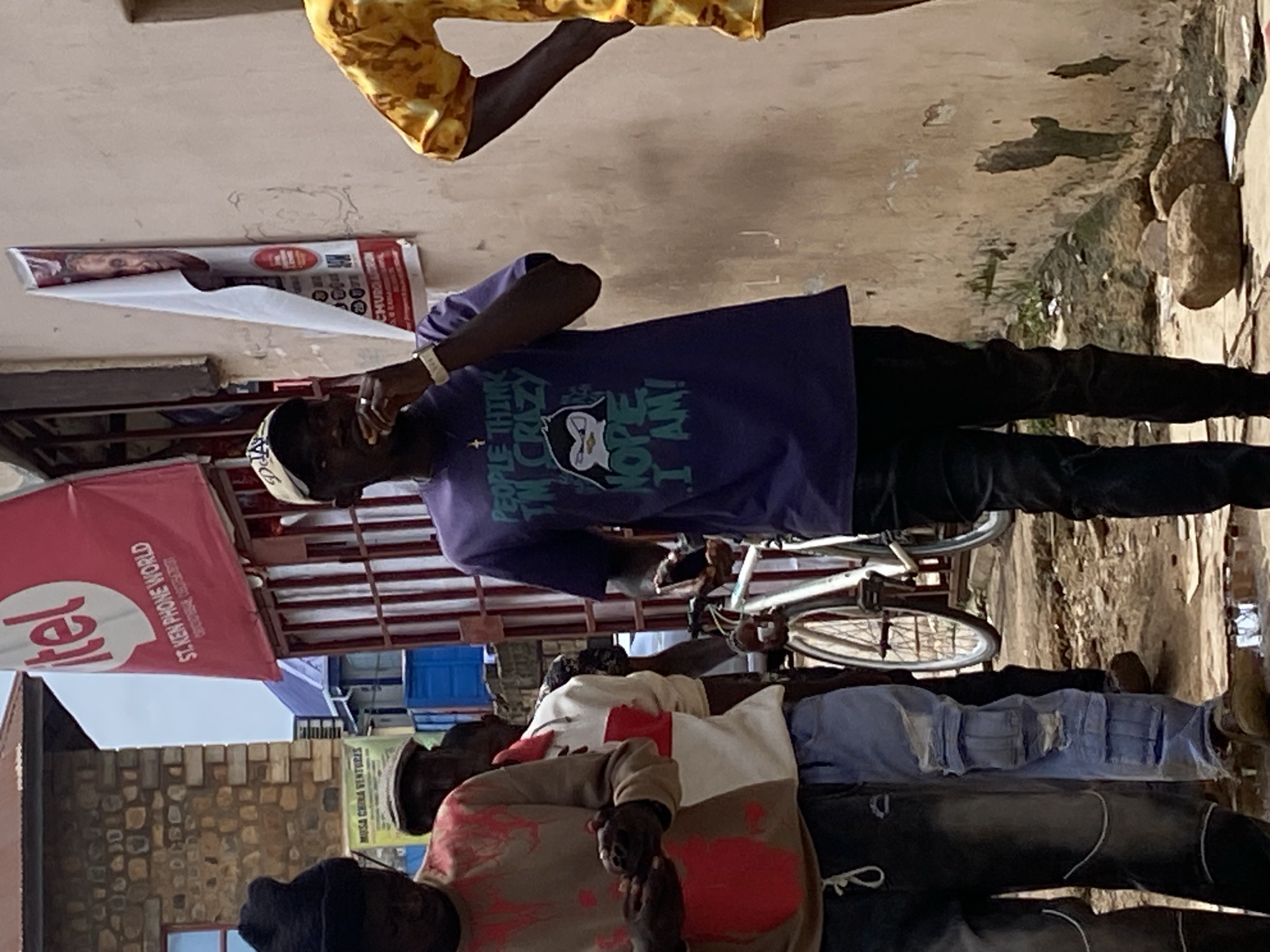 In an outdoor urban setting, several individuals are observed standing. One man in the foreground wears a purple t-shirt with the graphic "PEOPLE THINK I'M CRAZY NOPE I AM!". He is holding a small, dark object to his mouth, possibly food or a cigarette. To his left, another man in a tan sweatshirt with red paint splatters is looking down at his hands, which are holding a dark smartphone. He is wearing dark pants and a black beanie. Behind these two, a person in a white and red jacket is visible, and further behind them, another individual in a yellow and orange patterned shirt is partially visible. A white bicycle with a black seat and handlebars is parked to the left of the main subjects. In the background, a building with stone walls and a red sign that reads "itel" is prominent, along with a white banner displaying a man's face and red text that appears to be political or religious in nature. The ground is uneven, with dirt, debris, and scattered rocks. The wall to the right of the scene is weathered and has patches of peeling paint.