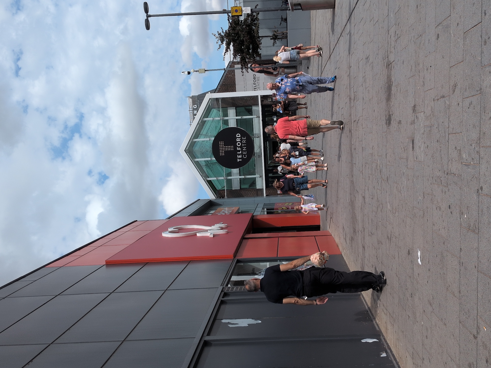 In a daytime outdoor scene in Telford, United Kingdom, people are walking along a paved pedestrian area in front of a modern building with a large red section featuring a white heart logo. The building on the left has a dark gray facade with vertical paneling. To the right of the red section, there is a glass-fronted entrance to a building with a large circular sign that reads "TELFORD CENTRE". Several individuals are visible walking in various directions. In the foreground, a man in a black polo shirt and black trousers is walking away from the camera, carrying a light-colored bag. Further down the pedestrian area, a woman in a blue shirt and blue trousers is walking towards the camera, accompanied by another woman in a floral dress and a man in a red shirt and shorts. A young child in a white dress is walking with the woman in the floral dress. Other individuals and a young child are also visible in the middle ground and background. The sky above is partly cloudy. A lamppost is visible on the right side of the image.