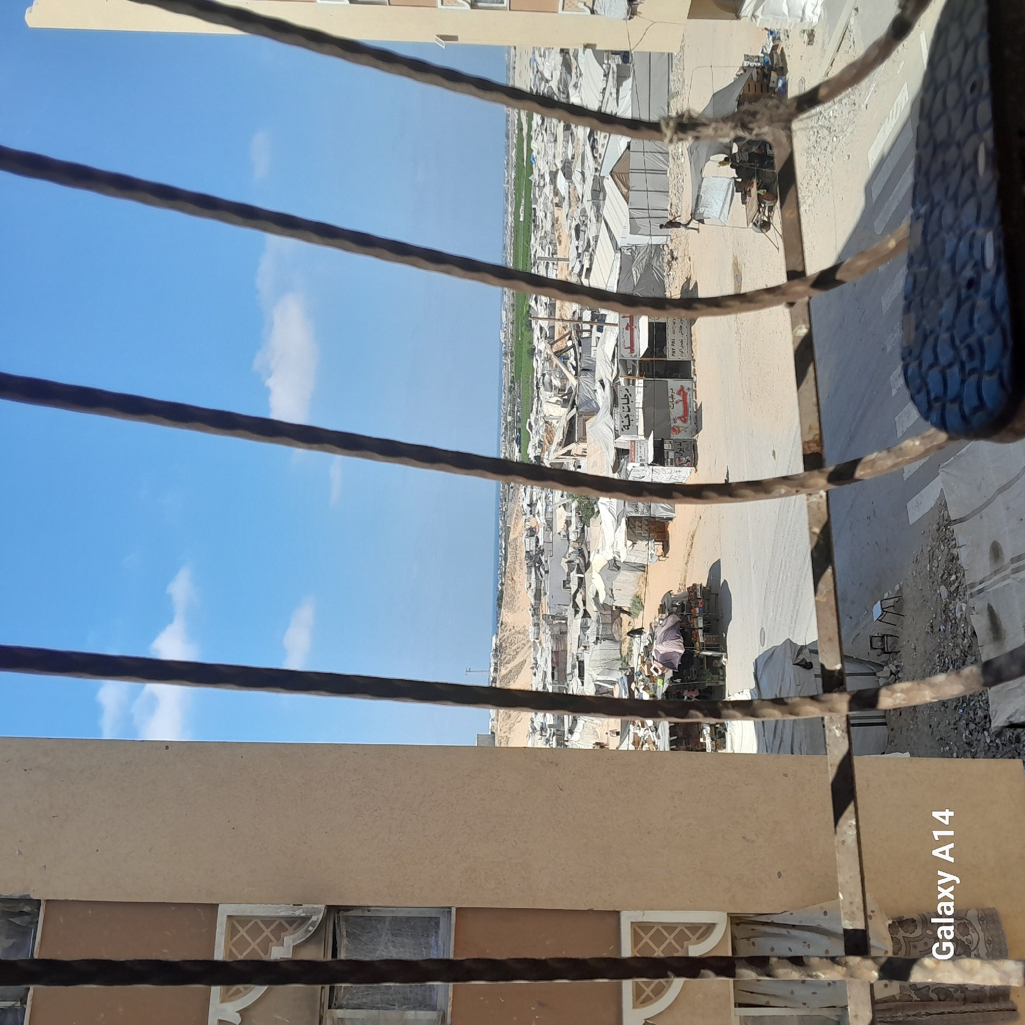 The image depicts an outdoor scene in the city of Khan Yunis, Palestinian Territories. The foreground is dominated by dark, weathered metal bars, suggesting a view from a window or a fence. Beyond the bars, a sprawling settlement of tents and makeshift structures is visible, characteristic of a refugee camp or informal housing. The tents are mostly white or light-colored, with some appearing damaged or in disrepair.
The environment is arid and dusty, with sandy ground and scattered debris. In the mid-ground, a dirt road runs through the settlement, with a few people visible walking or engaged in activities. There are also several small stalls or shops with awnings, some displaying signage with Arabic text. A small cart or vehicle with what appears to be a generator or engine is parked near one of the stalls.
Further in the background, a larger building, possibly part of a more permanent structure, stands on the right side of the frame. Beyond the settlement, a strip of green vegetation can be seen, contrasting with the arid landscape. The horizon shows the sea in the distance under a clear blue sky with scattered white clouds. The overall impression is one of a densely populated area with limited infrastructure, likely a community facing challenging living conditions. A watermark in the bottom left corner reads "Galaxy A14".