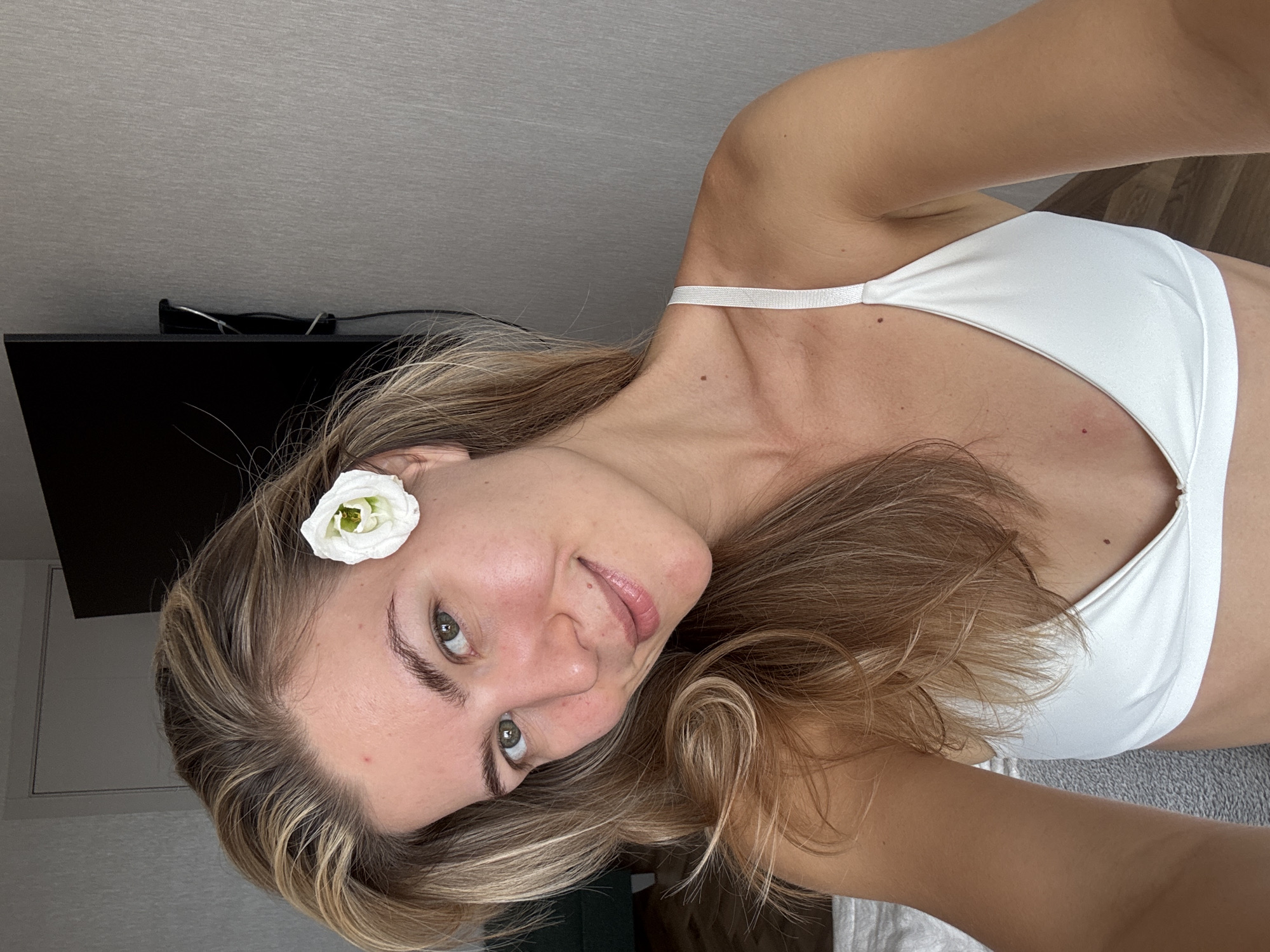 A woman with fair skin and blonde hair is positioned at an angle facing the camera. A white flower is situated above her ear. She is wearing a white top. In the background, there is a wall, a black object, and a wooden floor. The location is unspecified.