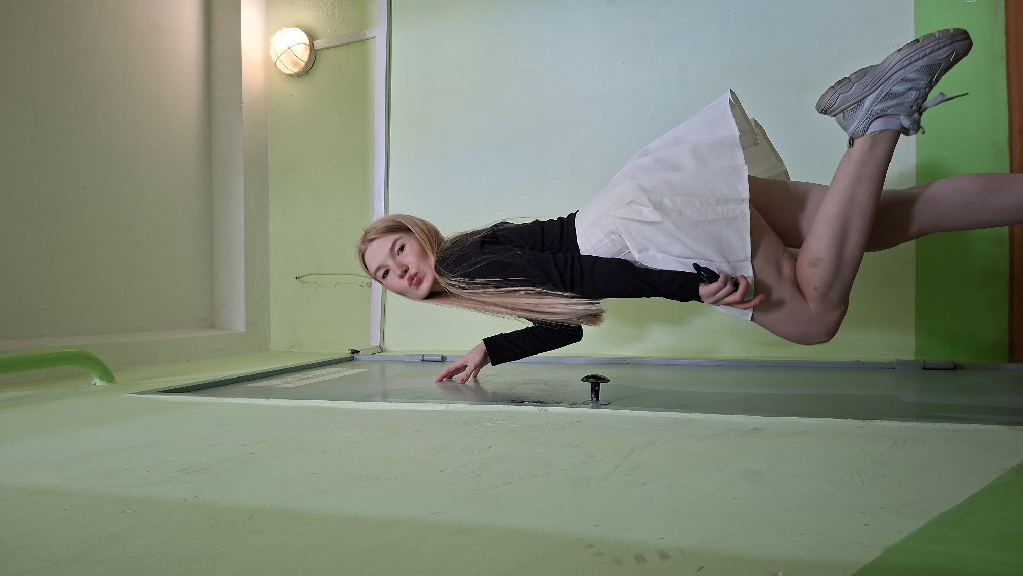 A young, blonde woman with fair skin poses in a hallway. She is wearing a black long-sleeved shirt and a white pleated skirt. Her left leg is bent and raised behind her, with her right foot flat on the floor. Her left hand is resting on a light green door, and she is puckering her lips towards the camera. The hallway walls are painted light green, with a white ceiling and a white light fixture on the right side of the image. A green pipe runs along the top of the left wall. The floor appears to be wood. The overall impression is one of a casual pose in a somewhat utilitarian indoor setting, likely a corridor in a residential building or school in Chaikovsky, Russia.