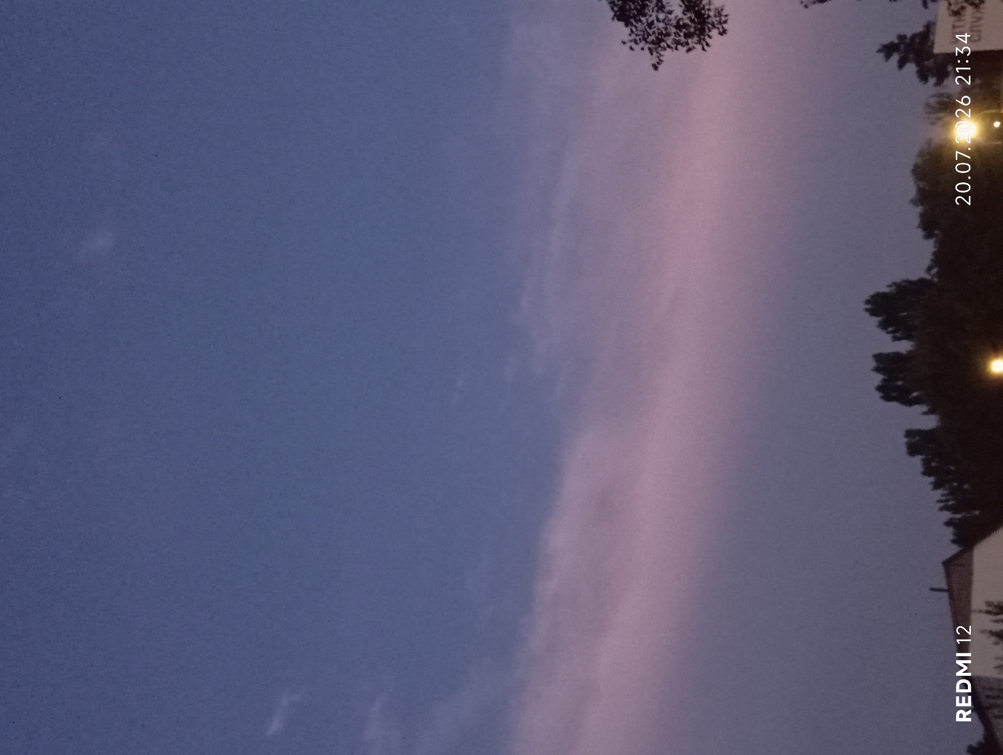 The image captures a twilight sky with a band of soft pink and purple clouds streaking across the otherwise deep blue expanse. Silhouetted trees and a building are visible along the right edge, suggesting an outdoor setting at dusk. The lighting indicates it is early evening, with the sky transitioning from day to night.
Visible text includes a timestamp "20.07.2026 21:34" and "REDMI 12" in white lettering, indicating the date and time the photograph was taken, as well as the model of the phone used. The location context provided is Smila, Ukraine. There are no people or active events depicted, the focus is on the atmospheric sky and surrounding landscape at the end of the day.