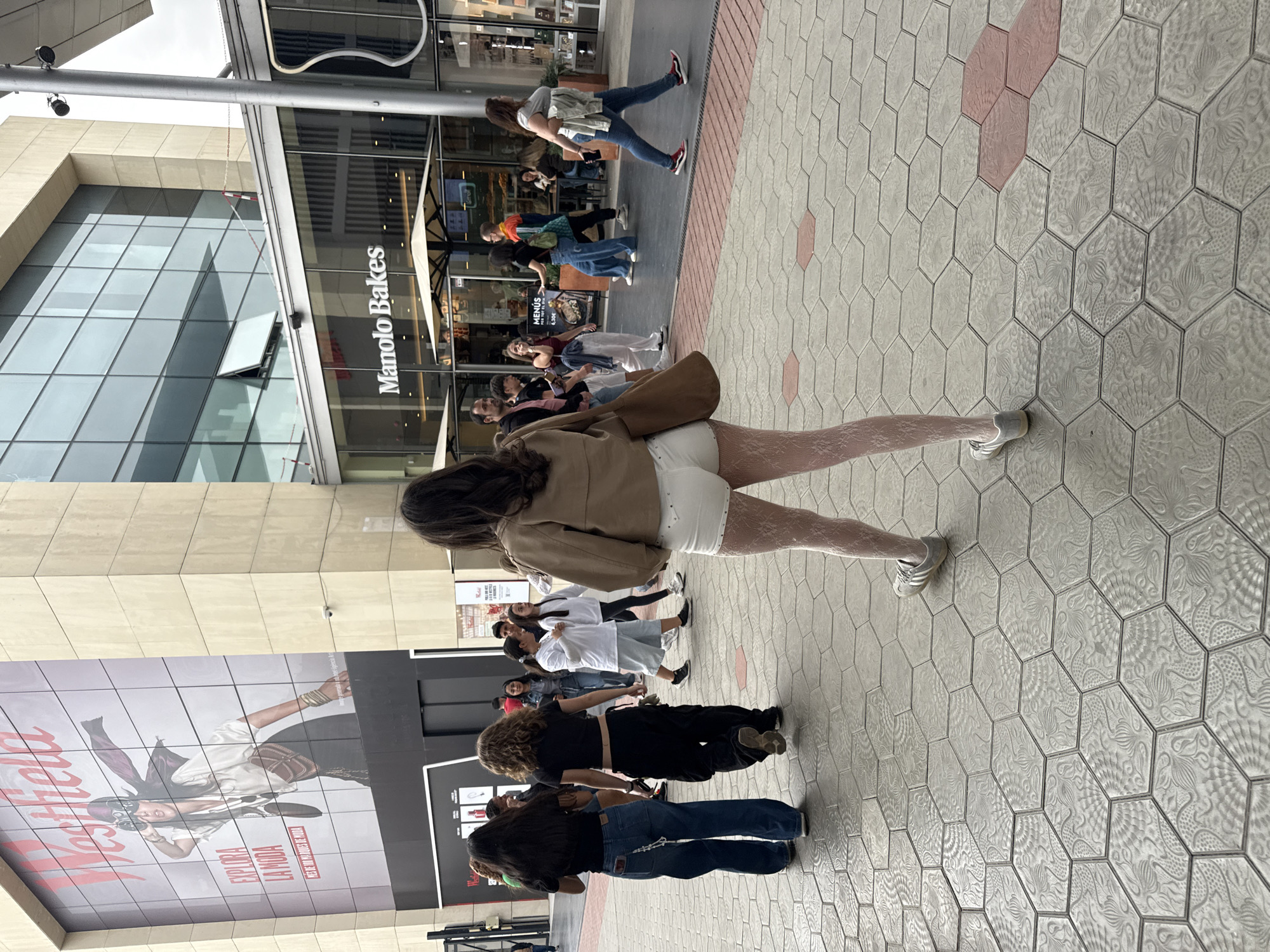 A medium, vertically oriented photograph shows a street scene with people walking and standing in front of a building with the words "Manolo Bakes" on the facade. The location is Westfield La Maquinista, Barcelona, Spain. The ground is paved with light grey hexagonal tiles with a decorative pattern, interspersed with light red hexagonal tiles. In the foreground, a woman wearing white shorts, patterned light pink tights, a tan jacket, and grey sneakers walks away from the camera. Several other people are walking or standing nearby, with varied clothing styles, including jeans, t-shirts, and dresses. A large billboard with the word "Westfield" and the phrase "EXPLORA LA MODA" is visible on the left. Behind the woman in the foreground is the glass front of "Manolo Bakes," and people are visible inside and outside. Above the storefront, the building has a modern architectural design with glass and metal elements. The sky is overcast.