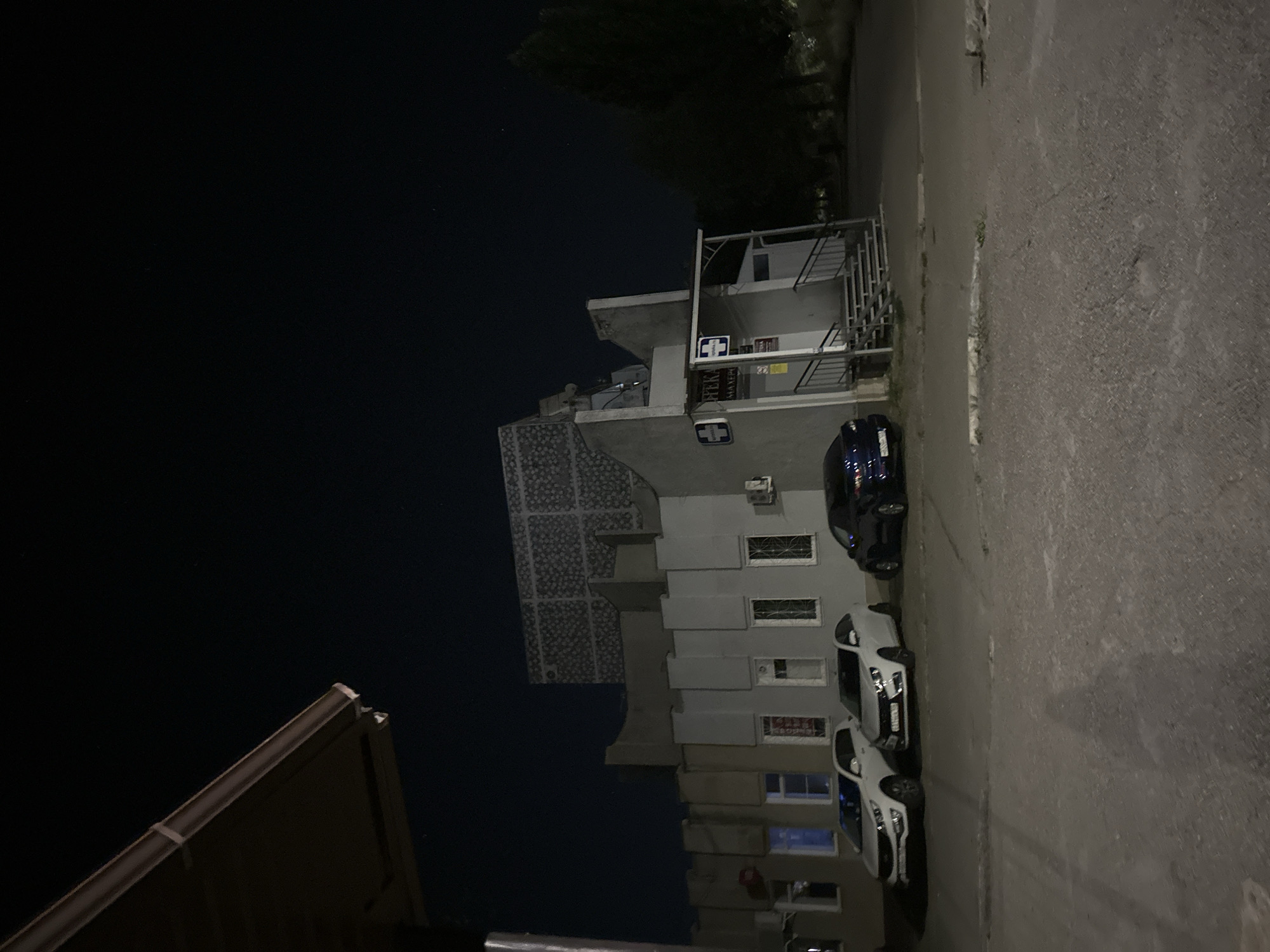 The image depicts a street scene at night in Nevinnomyssk, Russia. In the foreground, a dark asphalt surface is visible, with some gravel and debris. Three cars are parked: a white SUV on the left, a white sedan in the middle, and a dark sedan on the right. Behind the cars, a building with a grey facade is illuminated. The building has multiple windows, some with bars, and an entrance with stairs leading up to it. A sign with a white cross and text in a foreign language is mounted above the entrance. Further back, another structure with a distinctive patterned facade rises behind the main building. The sky above is dark, with a few faint stars visible. A dark tree line is present to the right of the building. A portion of a dark overhang or awning is visible in the upper left corner of the image. The scene appears to be a commercial or residential area.