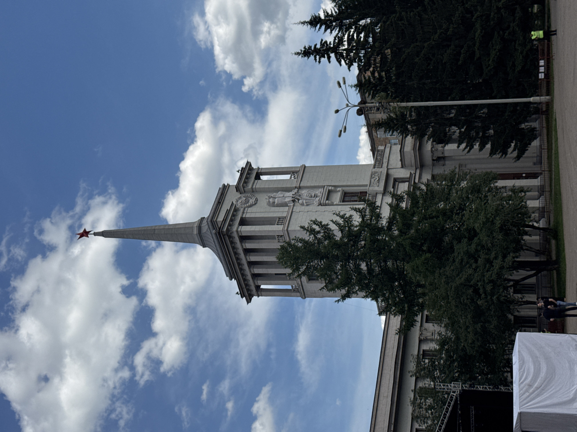The image shows the side view of a grand, ornate building with a tall, pointed spire topped by a red star. The building is constructed of light-colored stone and features classical architectural elements like columns and decorative reliefs. A large evergreen tree partially obscures the building on the right side. In the background, more trees and another building are visible. The sky is a vibrant blue with scattered white clouds, suggesting fair weather and daytime.
In the foreground on the right, a large white tent or structure, possibly a stage covering, is visible. Further to the right, two individuals are walking away from the camera on a paved pathway. One person is wearing a bright yellow safety vest, and the other appears to be in casual clothing. A metal truss structure, likely part of a stage setup, is also visible behind the white covering.
The overall scene appears to be an outdoor public space, possibly a square or park adjacent to the building. The presence of the stage equipment might indicate an event or performance is planned or has recently concluded. The architectural style and the red star on the spire are strong indicators of a location in Russia. The time of day is clearly daytime, and the weather is pleasant, with good visibility and a bright sky. No visible text is present in the image.
