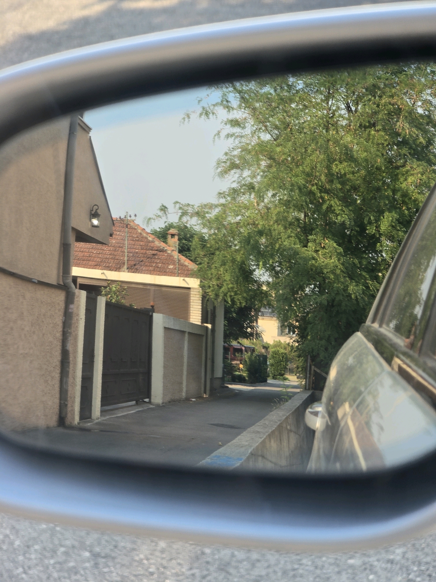 The image, taken from the perspective of a car's side-view mirror, shows a street scene in Podgorica, Montenegro. The mirror reflects a narrow street bordered by a light-colored building on the left and a wall with a dark gate. Further down the street, a red-tiled roof with a chimney is visible, as well as more buildings and dense green foliage from a large tree. The sky is clear and pale blue. The reflection also captures a portion of the car the mirror is attached to, appearing as a silver object in the right side of the frame. The road surface in the foreground and background of the mirror is asphalt, with some light and shadow patterns visible.