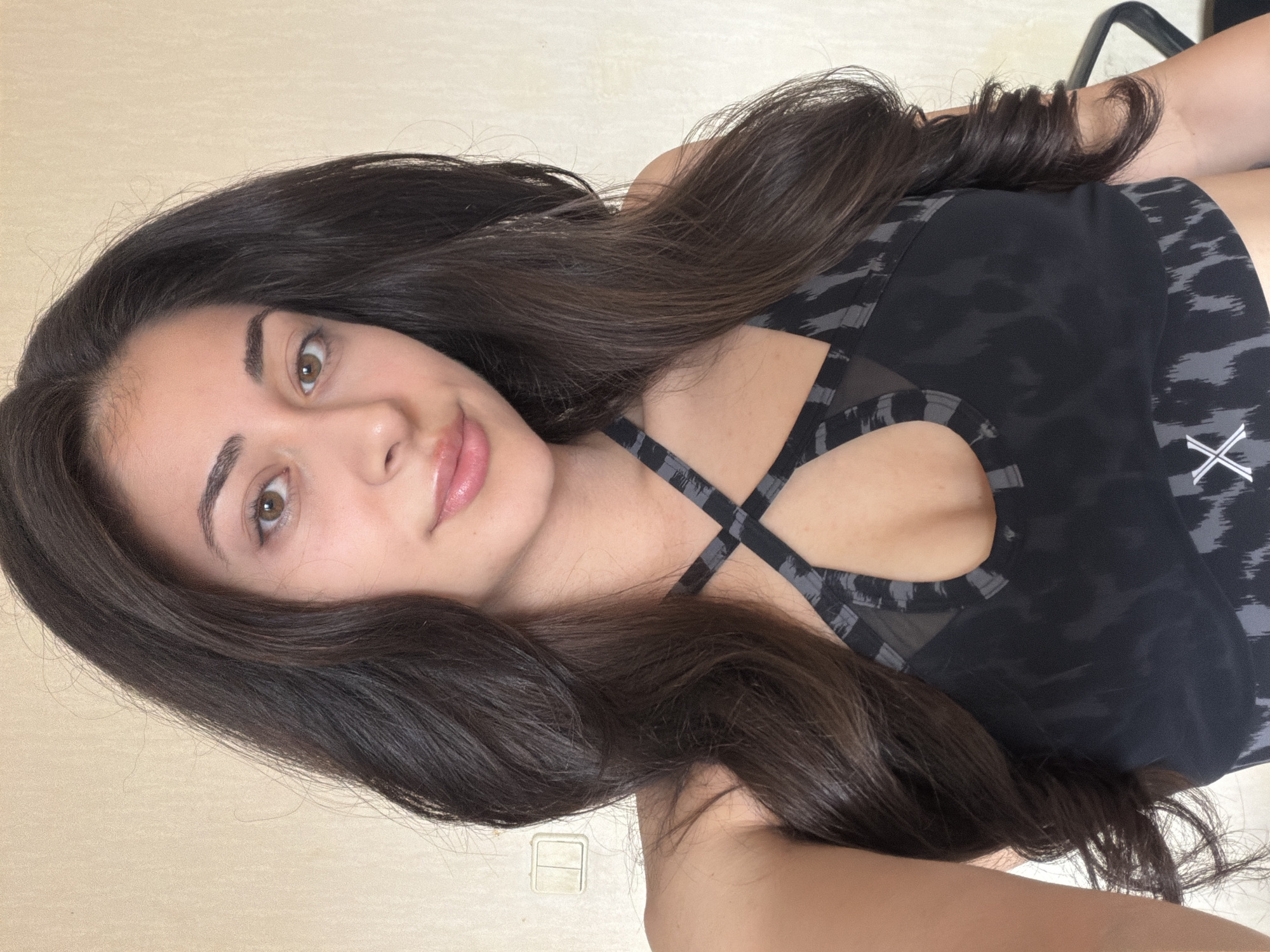 A young woman with long, dark, wavy hair and light brown eyes is depicted in a front-facing, chest-up shot. She is wearing a dark grey and black patterned sports bra with criss-cross straps across the chest, a central keyhole cutout, and mesh paneling. A white "X" logo is visible on the lower left of her garment. She has a neutral expression with a slight smile and is looking directly at the viewer. The background consists of a plain, light beige wall. On the left side of the frame, a cream-colored double rocker light switch is mounted on the wall. A dark, indistinct object, possibly a furniture piece, is partially visible on the far right. The scene is an indoor setting within the city of Sofia, Bulgaria.
