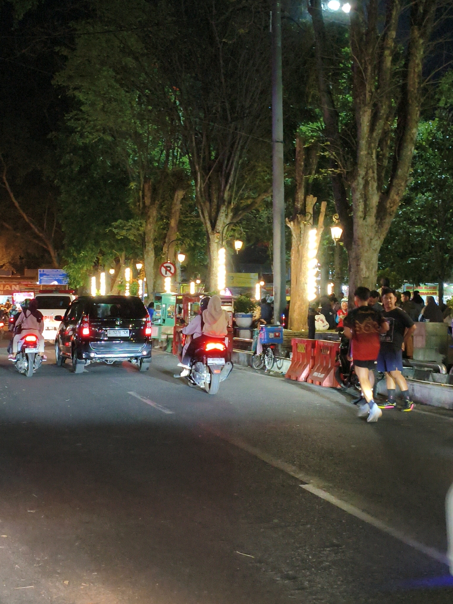 This is a vertical, eye-level, full-shot photograph taken at night on a city street in Salatiga, Indonesia. The scene depicts a moderate flow of traffic on a road lined with trees and illuminated by streetlights and decorative lights. In the foreground, a black car is moving away from the viewer, its taillights illuminated. To the left of the car, two motorcycles are also visible, one moving and one stationary. The motorcycle on the left has a rider wearing a light brown hijab and a light-colored outfit. The motorcycle to the right of the black car has a rider wearing a grey hijab and a dark outfit, with another person sitting behind them wearing a light brown hijab and a light-colored outfit. On the right side of the road, there is a sidewalk area with several people standing and walking. Two young men are prominent in the mid-right, one in a red shirt and dark shorts, the other in a black shirt and dark shorts. They are both looking towards the camera. Further back on the sidewalk, there are more people, some sitting and some standing, and several food stalls or small businesses are visible. The environment is urban, with trees and streetlights creating a canopy effect over the street. The overall impression is a lively evening scene in a commercial or public area of Salatiga.