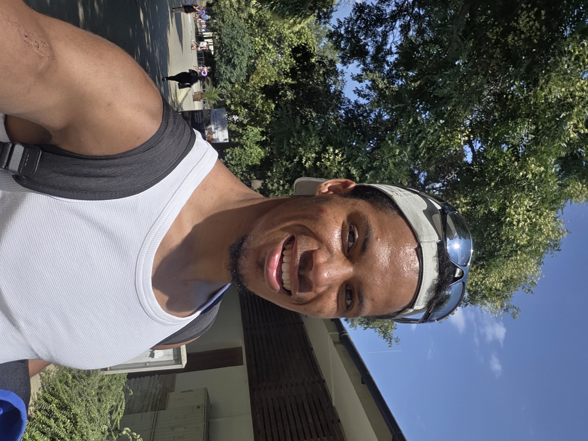 A man wearing a white tank top and a baseball cap with sunglasses on top of it is taking a selfie outdoors. He has a beard and is smiling. In the background, there are trees, a building with wooden slats, and several people walking on a path. The scene appears to be a sunny day in Varna, Bulgaria, possibly near the Sea Garden.