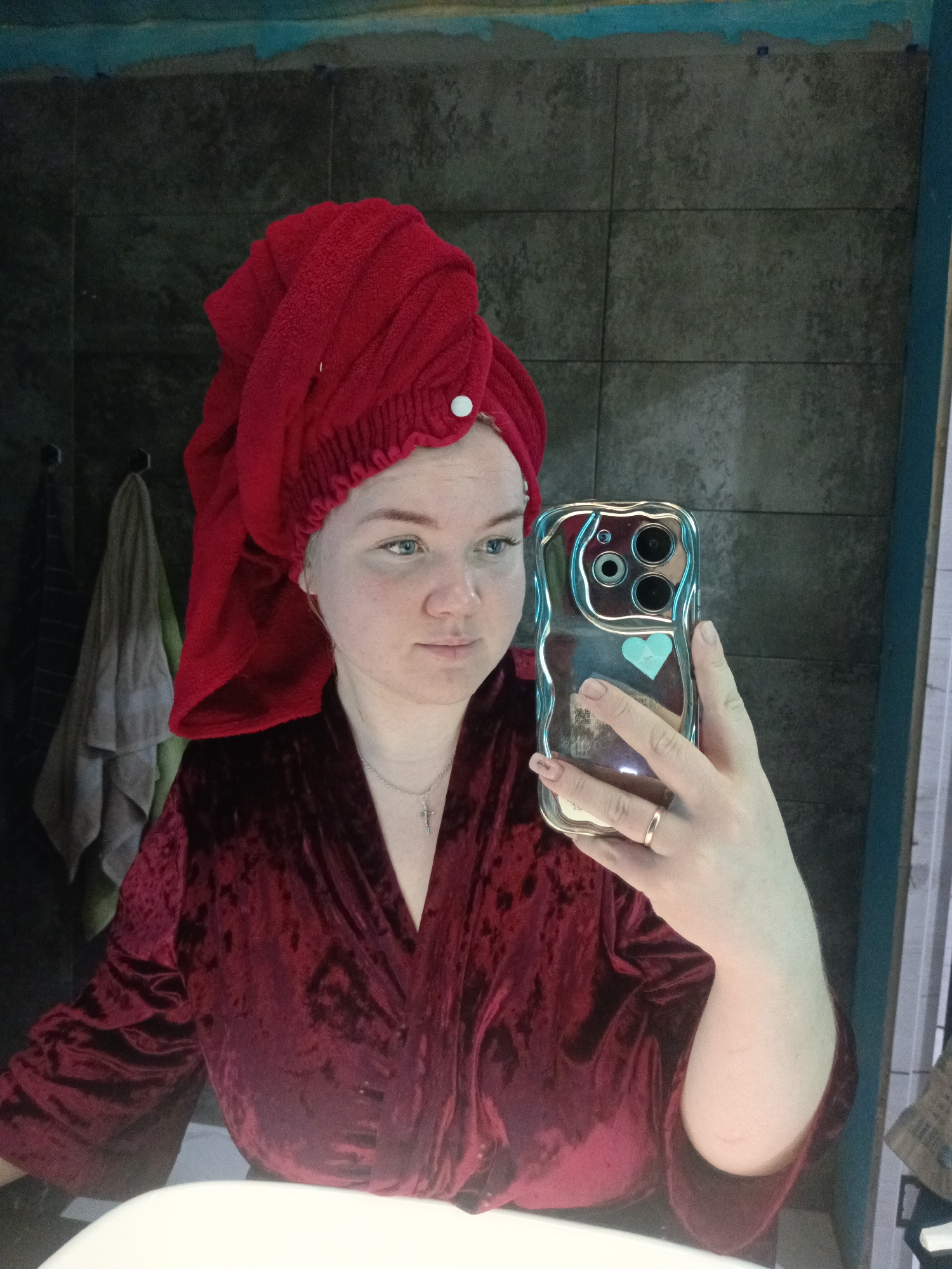 A person wearing a burgundy velvet robe and a red towel wrapped around their head is taking a selfie in a bathroom. The person is holding a phone with a silver case. The background consists of gray tiled walls and a glimpse of hanging towels. The location, Фоки, Russia, cannot be determined from this image alone.
