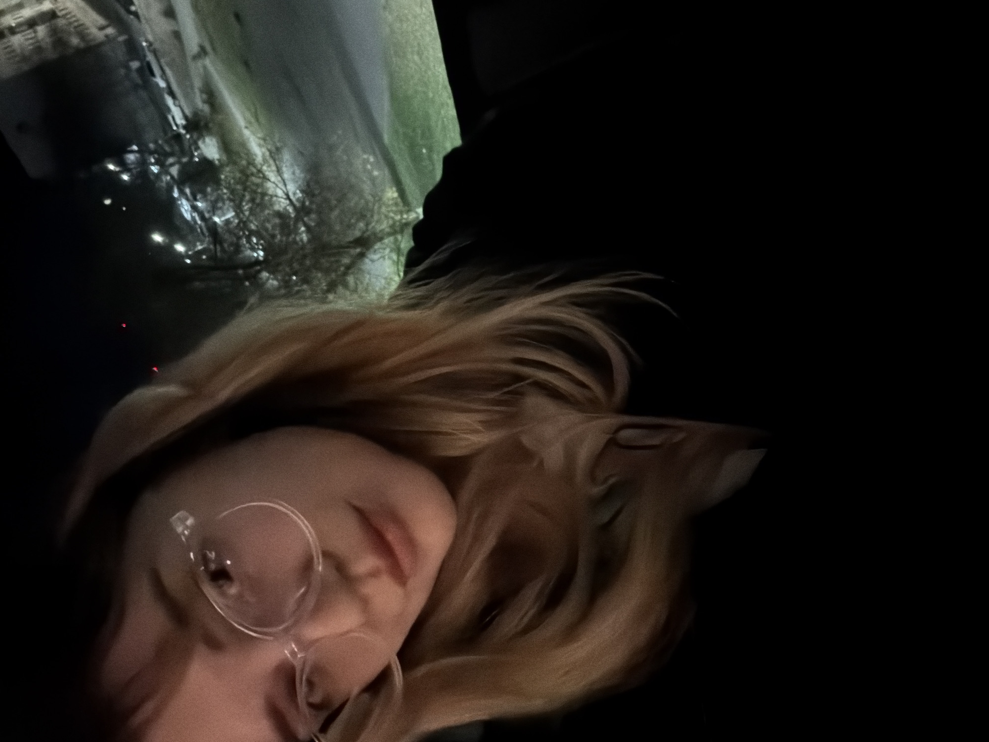 A nighttime view shows a person with blonde hair and glasses, possibly female, facing the camera in a selfie style. The background reveals an urban environment. Based on context, it seems this photo may have been taken in Naberezhnye Chelny, Russia, based on the presence of buildings and streetlights that align with images from the city. There is limited light in the scene, with the primary sources being street lighting reflected on nearby buildings.