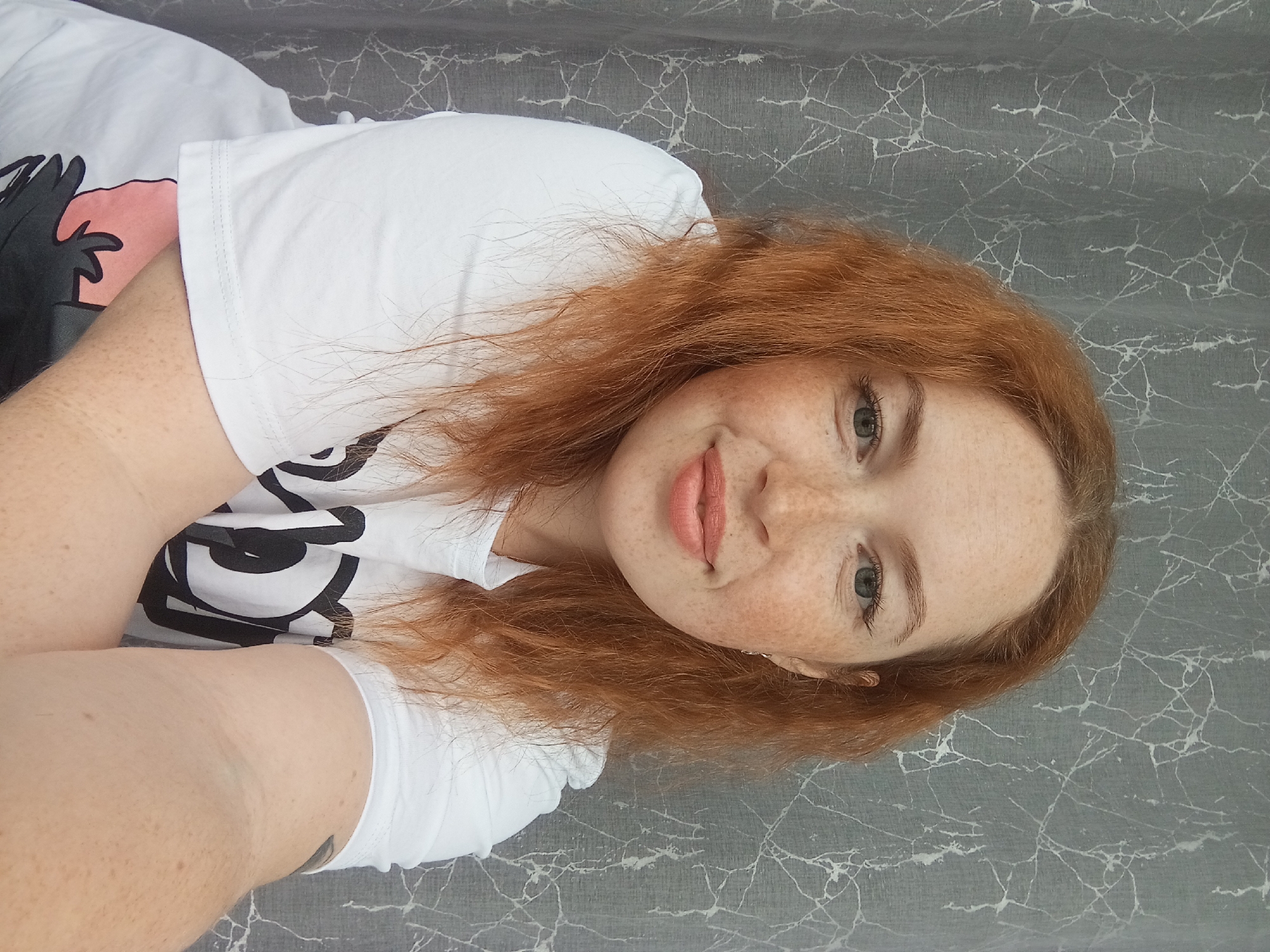 A young woman with red hair and freckles is lying on a gray surface with a white marble-like pattern. She is wearing a white t-shirt with a black and white graphic design. Her eyes are light-colored, and she has a slight smile. The location is potentially in Yurgamysh, Russia.