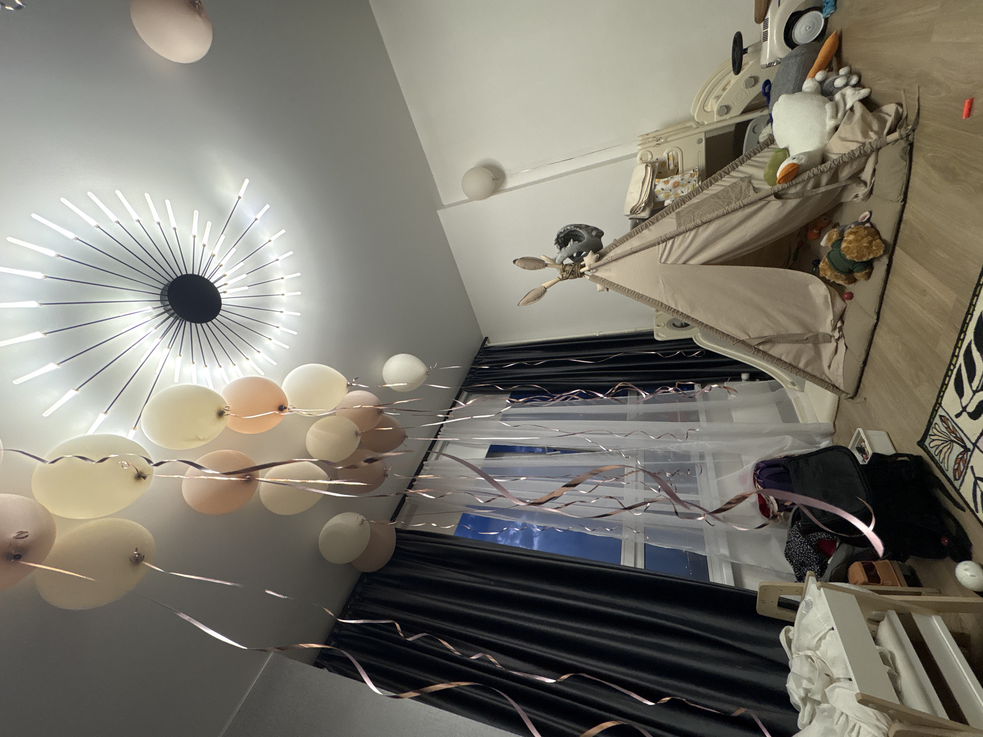 The interior scene shows a child's room, likely in Unknown, Unknown. A teepee-style tent is positioned in the corner of the room, filled with plush toys including a white goose, carrot, and teddy bear. Adjacent to the teepee is a baby activity center and other toys. Dark grey curtains frame a window, with a clear view of the sky visible through the glass. Several pastel-colored balloons with ribbons are suspended from the ceiling. Above, a modern, starburst-style ceiling light fixture illuminates the room. A baby crib with white linen is partially visible at the bottom of the frame, and various bags and containers are cluttered on the floor near the window.