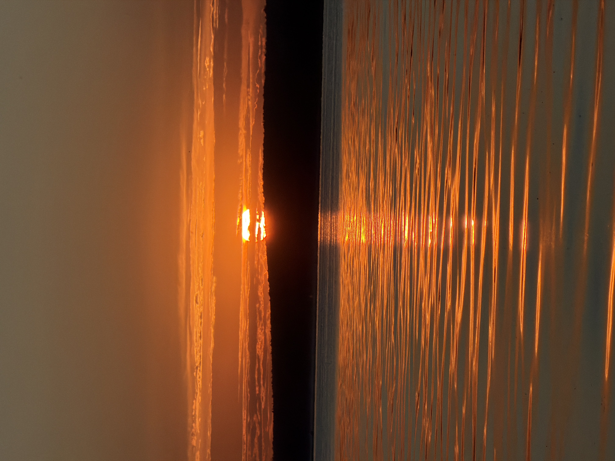 An image captures a serene sunset over a body of water, likely the Kama River near Chaikovsky, Russia. The sky is a gradient of warm hues, transitioning from a deep orange near the horizon to a muted brown at the top. Wispy clouds are scattered across the sky, tinged with orange by the setting sun. The sun itself is partially obscured by a dark, silhouetted treeline that stretches across the middle ground, creating a stark contrast against the bright sky. The treeline appears to be the edge of a forest or a hilly landscape.
The foreground is dominated by the calm surface of the water, which reflects the vibrant colors of the sunset. Gentle ripples on the water create horizontal lines of light and shadow, enhancing the visual texture. The reflection of the sun appears as a shimmering pathway of orange light stretching across the water towards the viewer. No people, specific objects, or actions are discernable in the scene. The environment appears natural and tranquil, typical of a sunset view over a river or large lake.
