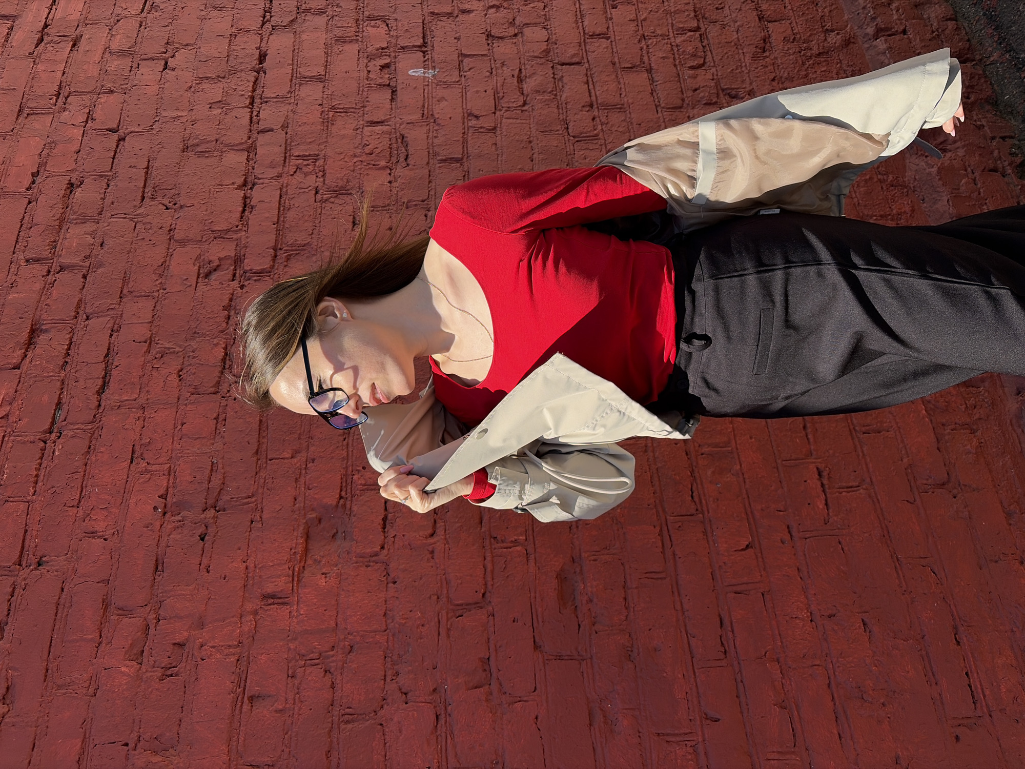 A young woman with light brown hair and eyeglasses is lying on a red brick surface. She is wearing a red top, black pants, and a tan-colored trench coat. The image is oriented so that the woman appears to be sideways. The background consists entirely of the red brick surface. The location is potentially in Izhevsk, Russia.