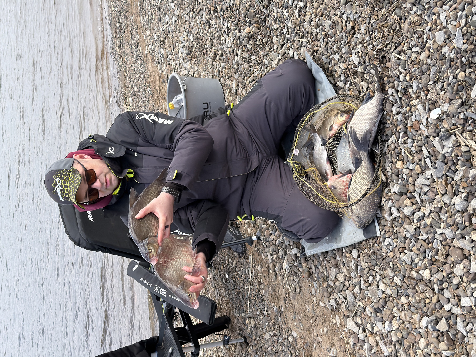 This image captures a fisherman seated on a rocky shore beside a calm body of water, likely a river or lake, in Chaykovsky, Russia. The individual, wearing dark, heavy-duty fishing attire including a jacket with a "MAX" or "MARU" logo on the sleeve, dark trousers, sunglasses, and a grey cap with a yellow hexagonal pattern, is holding a freshly caught fish, resembling bream, in both hands.
To the fisherman's right, a yellow mesh keepnet is placed on the pebbles, brimming with several other fish, indicating a successful catch. A grey bucket with "MARU POWER" printed on it and a plastic bottle are positioned behind the fisherman. The black fishing chair he is sitting on features text including "JURA" and "FEEDER".
The setting features a shoreline covered in various shades of grey and brown pebbles. The water appears calm and grey, suggesting an overcast day rather than bright sunlight, although the fisherman is wearing sunglasses. The image is taken from a slightly elevated viewpoint, focusing on the fisherman and his substantial catch.