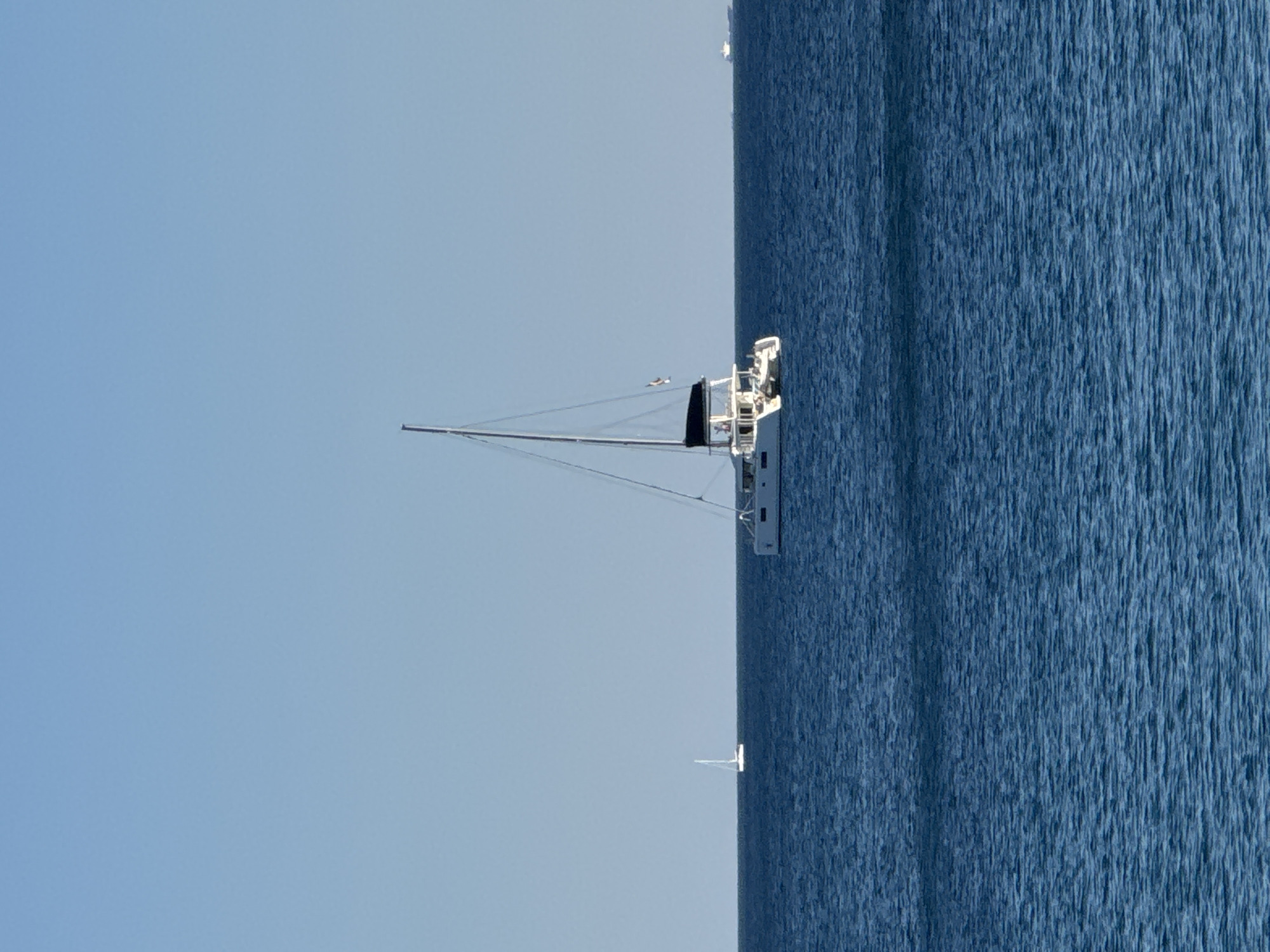 The image, oriented vertically, depicts a serene seascape. The scene is divided into two main sections: a vast expanse of clear, pale blue sky on the left and the deep, textured blue of the sea on the right. The sea exhibits a gentle ripple, suggesting a calm day.
In the foreground, a portion of a sailboat is visible. Its mast extends upwards, and rigging is apparent. The boat appears to be white with dark accents, and a white structure with railings is perched on its deck. Two small figures, possibly people, are visible on the deck of the sailboat.
Further in the distance, on the horizon where the sea meets the sky, another small white object, likely a distant boat or buoy, is faintly visible. There is no visible text in the image. The lighting suggests it is daytime, likely mid-morning or afternoon, with clear weather conditions. The overall mood is peaceful and tranquil, characteristic of a quiet day at sea. The location context provided indicates Moschato, Greece, which aligns with a coastal environment.