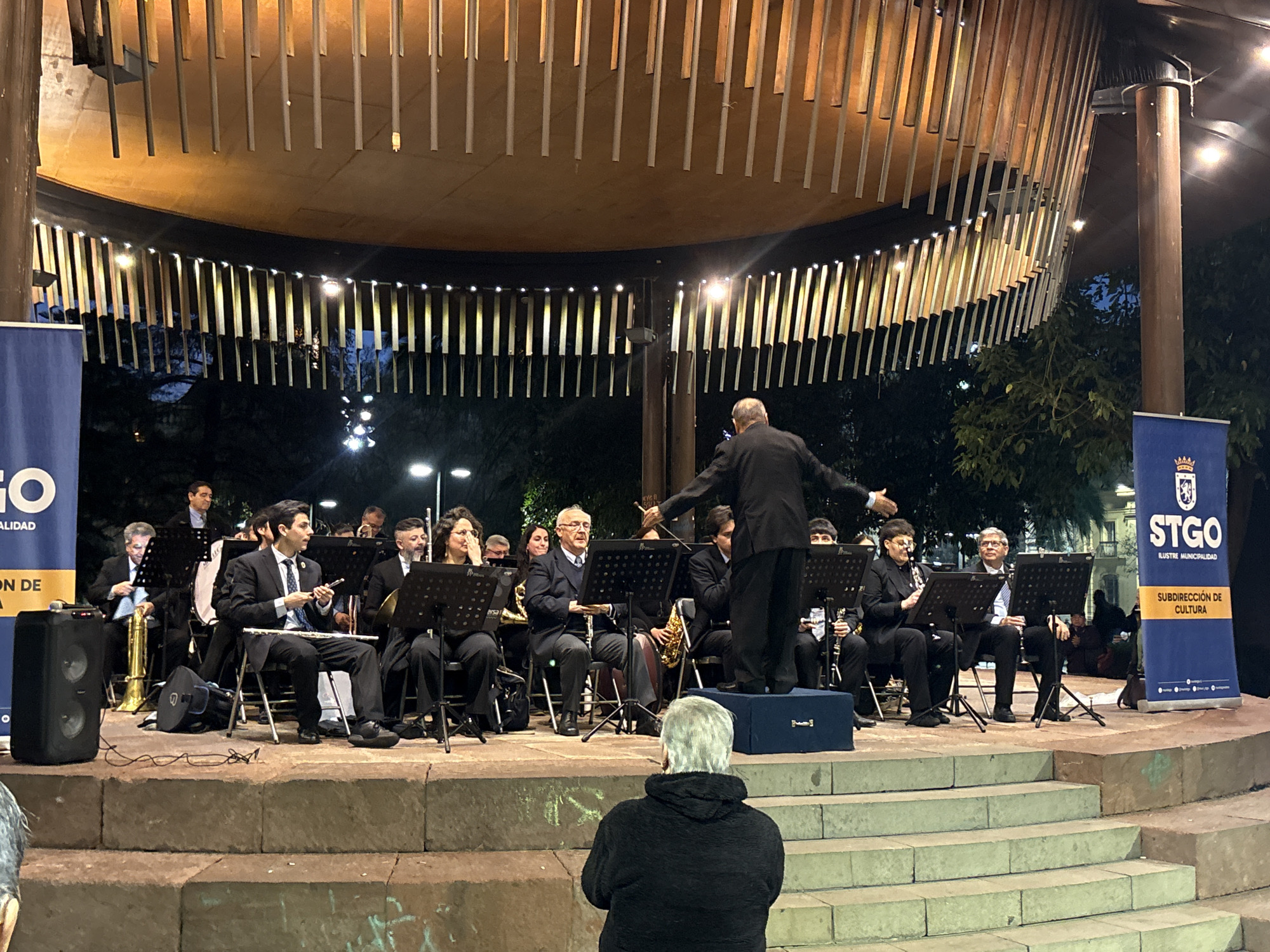 The image depicts an outdoor evening concert performance by an orchestra in an amphitheater-like structure, likely in the city of Santiago, Chile, indicated by the presence of banners that say "STGO ILUSTRE MUNICIPALIDAD" and "SUBDIRECCIÓN DE CULTURA". A conductor, facing away from the camera, leads the ensemble from a raised platform. The orchestra members, all dressed in formal attire, are seated and playing various instruments including clarinets, saxophones, and brass instruments. Music stands are positioned in front of each musician. In the foreground, facing the performers, sits an individual with gray hair, wearing a dark jacket, appearing to be an audience member. The stage area is illuminated by overhead lights and string lights decorating the structure's canopy. The background shows trees and further urban structures, suggesting a city park or public square setting.
