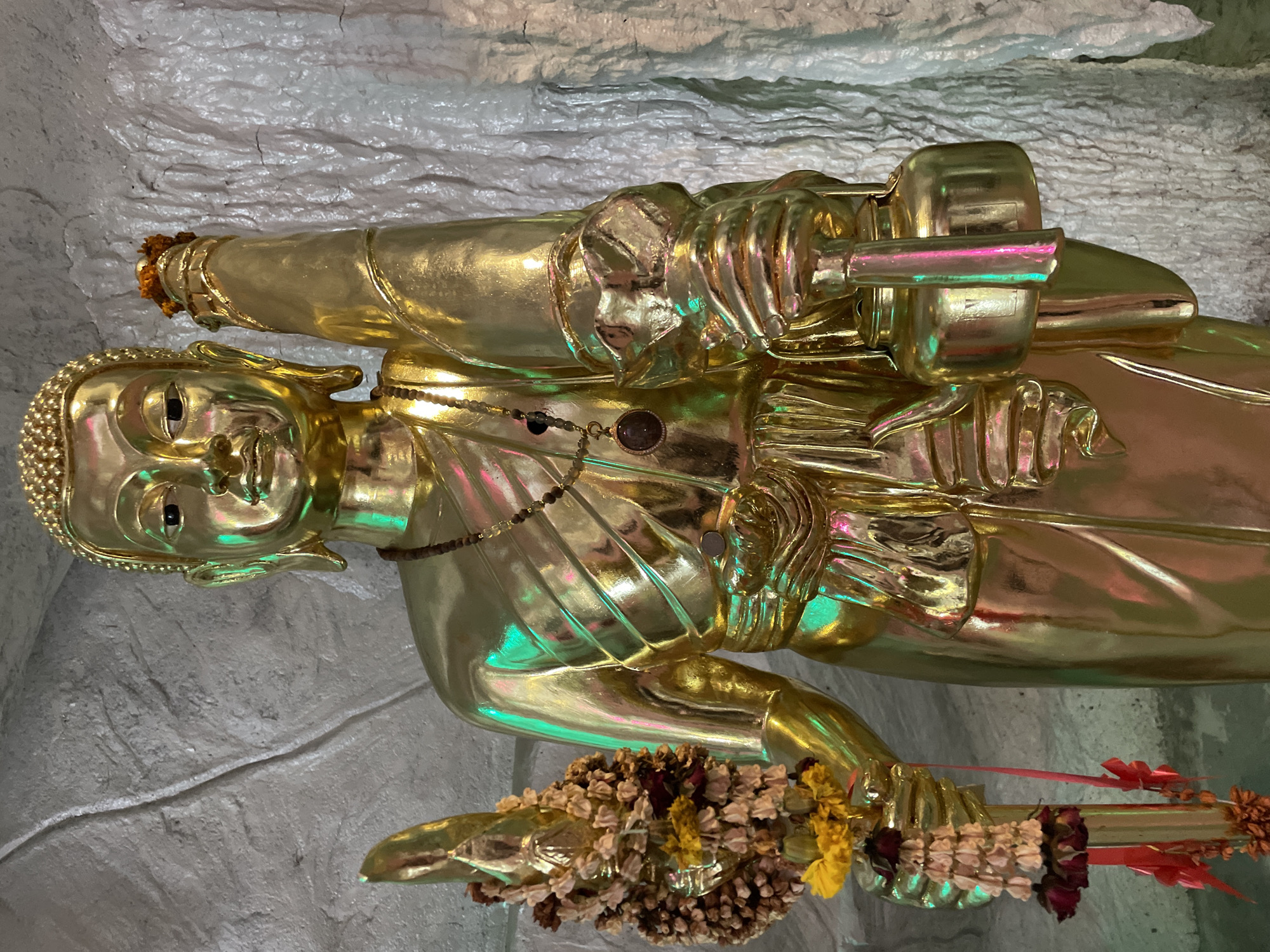 A golden statue of a monk stands in a cave-like environment. The statue is adorned with a beaded necklace and holds two objects. In its left hand, it grasps a staff topped with a floral offering, while its right hand holds a round object with a spout. The cave walls are textured and off-white. The scene is illuminated with colored lighting, casting green and pink reflections on the statue. The location is identified as Bo Win Subdistrict, Thailand.