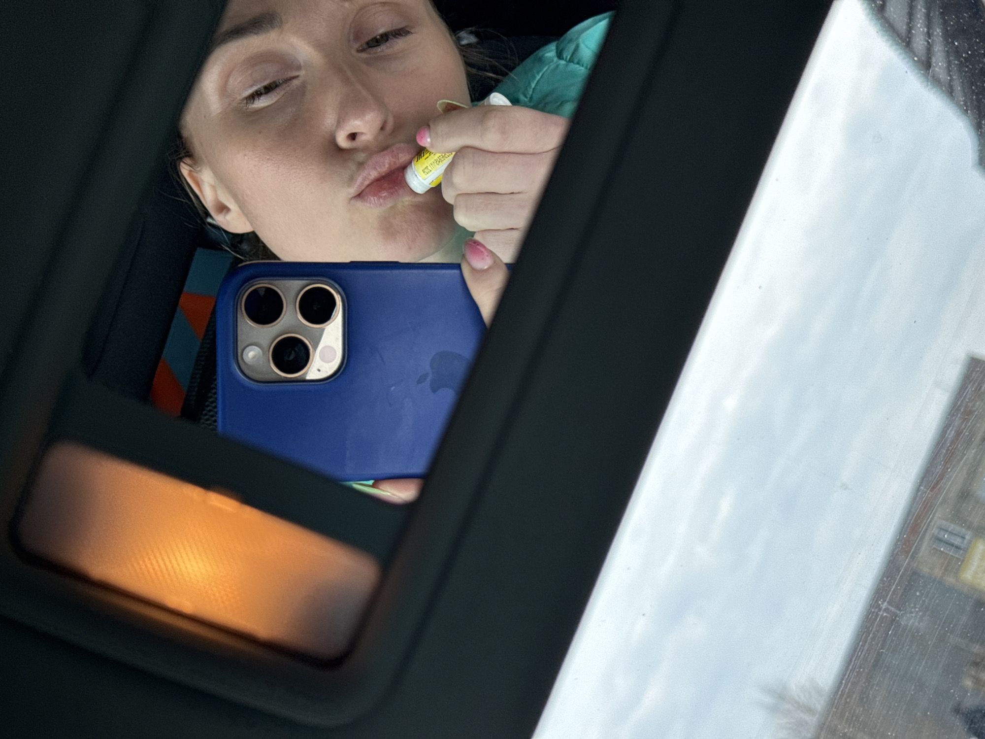 A woman is applying lip balm while looking at herself in the reflection of a blue-cased iPhone. The scene is viewed from inside a vehicle, possibly in Chaikovsky, Russia, as indicated by the background visible through the car's window. The car's sun visor is down.