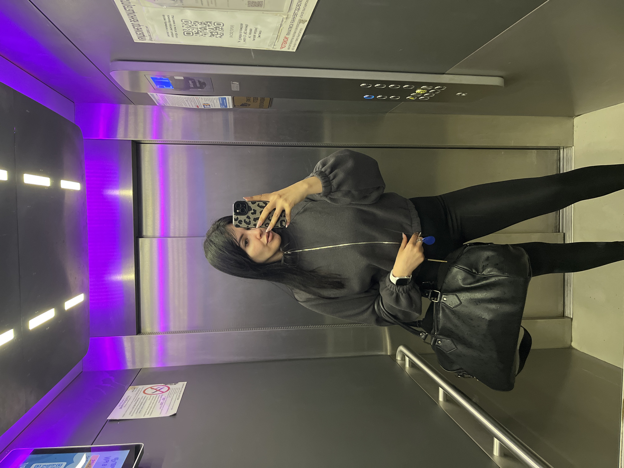 The image shows a woman taking a selfie in an elevator. She is wearing a grey zip-up sweater and black leggings. She is holding a black bag and a phone with a leopard print case. The elevator has purple lighting. There is a sign in the elevator in Russian, which is associated with the city of Sochi, Russia. The sign contains a QR code and other information. There is also a panel with buttons on the wall of the elevator.