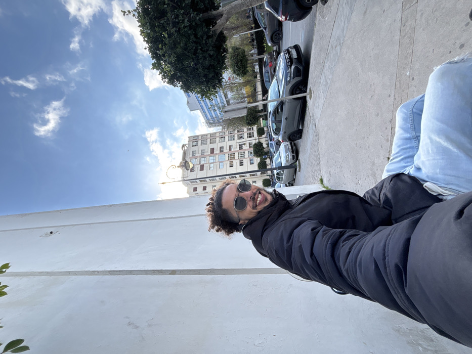 An outdoor urban scene in Tangier, Morocco, captured during daylight under a partly cloudy blue sky. A male subject, appearing to be in his 20s or 30s, is positioned in the left foreground, framed against a tall, plain white wall. He possesses dark, curly hair and stubble, wears dark-rimmed sunglasses, a black puffer jacket, and light blue jeans. His expression is a smile directed towards the camera, with his right arm extended, indicative of a selfie capture. The subject is situated on a concrete sidewalk. Beyond the sidewalk, a paved street contains multiple parked vehicles, including a silver sedan and a prominent grey Dacia Sandero Stepway. The background features several multi-story buildings: a light-colored structure with numerous windows and another modern building with a blue-tinted facade. An ornate black street lamp is visible, with the sun's glare originating from behind it. Green, leafy trees line the street on the right side of the frame.