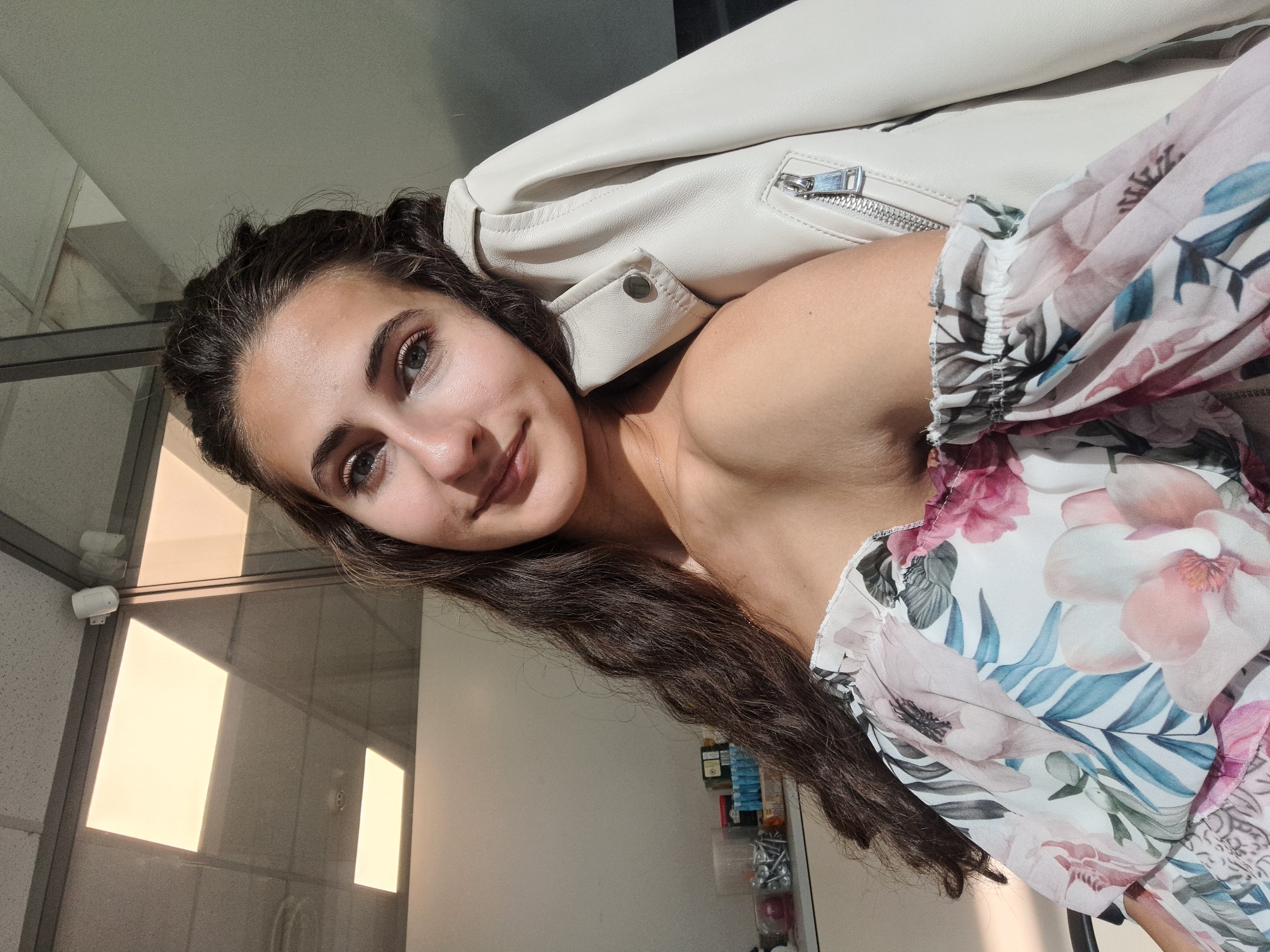 In an office environment with fluorescent lighting visible on the ceiling, a young woman is captured in a selfie. She has long, dark, wavy hair and is wearing an off-the-shoulder floral print top. A cream-colored leather jacket is draped over her shoulder. She is looking towards the camera with a slight smile. The background suggests an indoor setting with glass partitions and office furniture, hinting at a location within the city of Sofia, Bulgaria.