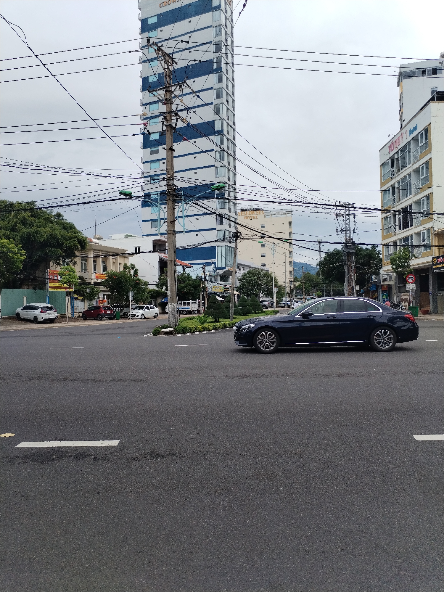 The image captures an urban scene in Nha Trang Ward, Vietnam. In the foreground, a black Mercedes-Benz sedan is driving across the frame on a multi-lane asphalt road. Road markings, including white dashes, are visible on the pavement.
Behind the car, the cityscape is dominated by several buildings. A tall, modern high-rise with white and blue horizontal stripes is prominent in the midground, partially obscured by a utility pole and a complex network of overhead wires. Adjacent to it is a shorter, light-colored building with windows and signage.
Further in the background, more buildings and trees can be seen, indicating a densely populated area. The sky is overcast, suggesting a cloudy day. Other parked cars are visible on the left side of the road.