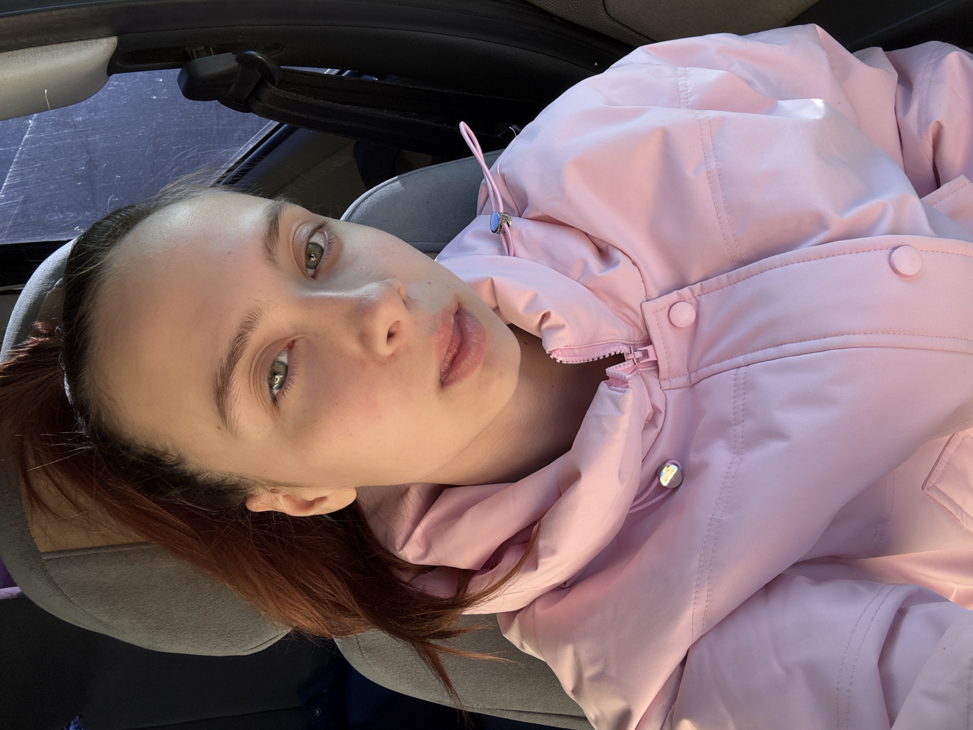 A close-up selfie captures a young woman inside a car, presented at an angle as if she is reclining. The woman has fair skin, light brown hair pulled back into a ponytail, and striking green eyes, maintaining a neutral gaze directly towards the camera. She is dressed in a light pink puffer jacket, which features a front zipper, decorative snaps, and drawstrings with metallic silver toggles near the collar.
The setting is clearly the interior of a vehicle, with parts of a grey car seat headrest and a dark ceiling visible in the background. A car window behind her shows a dark, somewhat scratched or textured surface outside, indicating the daytime. Natural light illuminates her face and jacket, creating bright highlights and soft shadows, suggesting either morning or late afternoon. The puffer jacket implies a cooler climate, fitting with the provided location context of Прикамский, Russia. The image focuses solely on the woman and her immediate surroundings within the car, with no other discernible activities or events taking place beyond posing for the photo. There is no visible text within the frame.