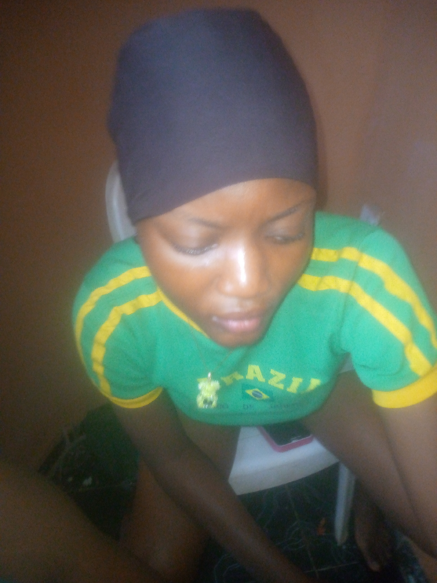 A woman wearing a dark head covering and a green and yellow t-shirt with the word "BRAZIL" and a Brazilian flag is sitting on a white plastic chair. A dark smartphone is placed on the chair near her. The photo was taken indoors against a wall with a brown tone. It is not possible to confirm the location as Ikorodu, Nigeria.
