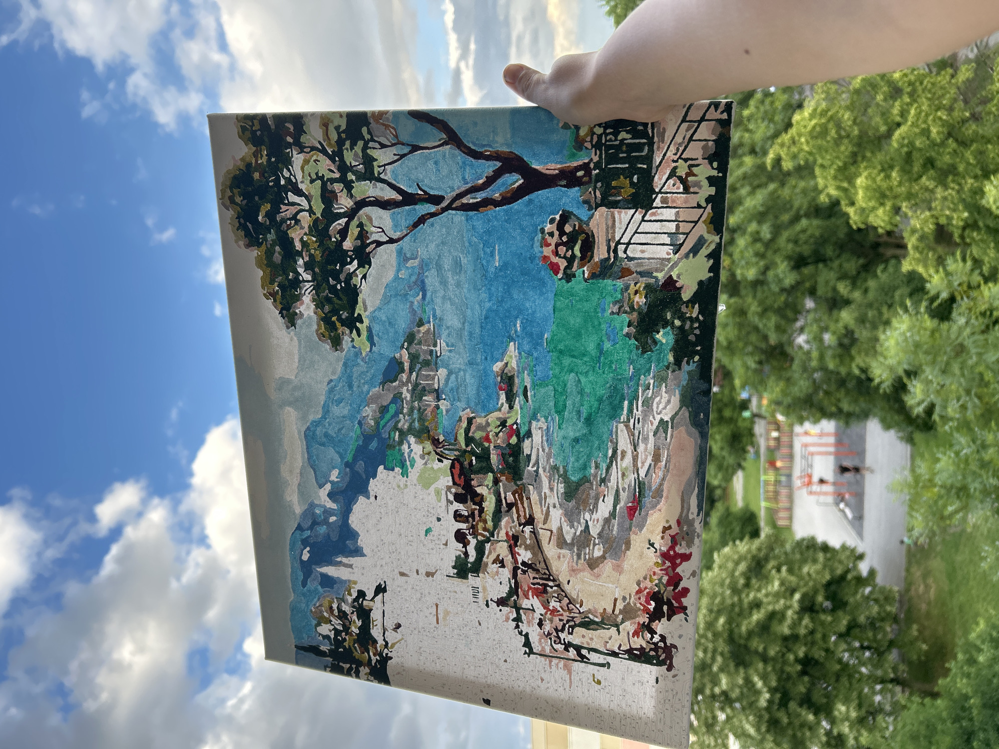 A partially completed paint-by-numbers canvas is held up by a person's hand, positioned against a vibrant blue sky filled with scattered white clouds. The canvas depicts a picturesque landscape, likely a Mediterranean coastal scene, featuring a large, dark-branched tree with green foliage, a decorative railing, and buildings descending towards a body of water in varying shades of blue and green. The top and right portions of the painting are mostly colored, showcasing a blend of green for vegetation, blues for water and sky, and earthy tones for structures. The lower and left sections of the canvas remain largely unpainted, revealing white areas marked with small numbers, indicating the in-progress nature of the artwork.
The setting appears to be outdoors, from an elevated viewpoint, possibly a balcony or window overlooking a green area. In the background, beyond the canvas, dense green treetops are visible, suggesting a park or courtyard. A close examination of the background in one of the provided crops reveals a blurred view of a playground with colorful structures and faint outlines of what appear to be people.
The scene is illuminated by bright daylight, indicating a sunny day with fair weather. The time of day is clearly daytime. No overt emotions are discernible. The only visible "text" consists of the small numbers on the unpainted sections of the paint-by-numbers canvas, guiding the artist. The overall location context is Sofia, Bulgaria.