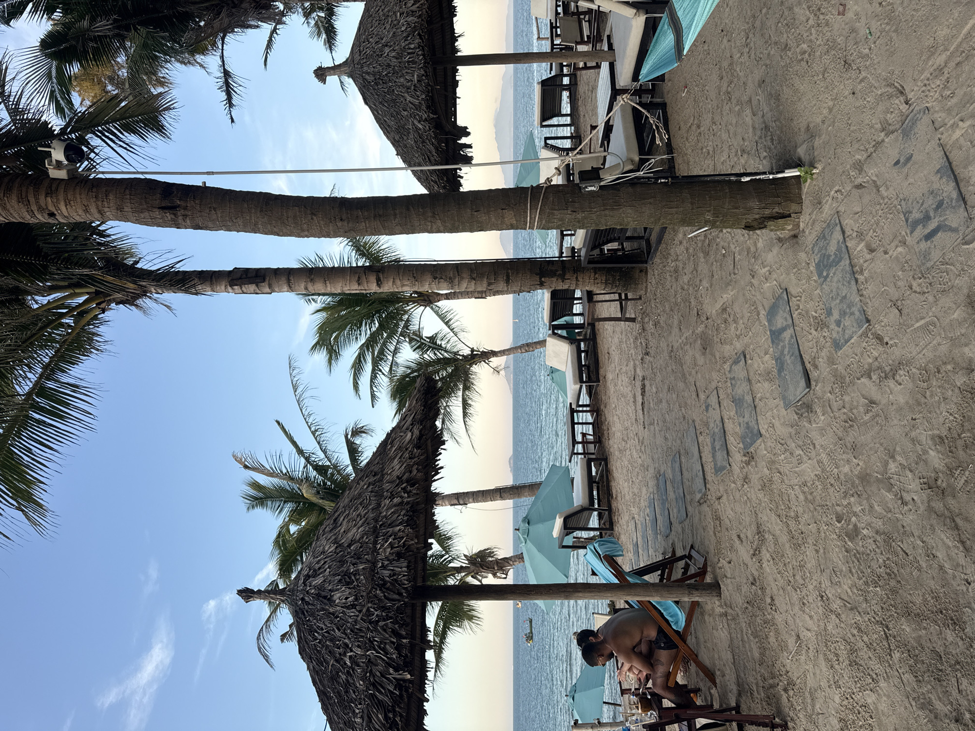The image depicts a beach scene with several elements:
**Environment:** A sandy beach with palm trees and straw parasols. The sky is light blue with some clouds. The ocean is visible in the background.
**Objects:** Beach chairs with blue towels, tables, umbrellas, and stone pathways are present.
**People:** There are two people sitting in beach chairs.
**Actions:** People are seated and appearing to engage in conversation or an activity.
**Location:** Unknown City, Unknown.