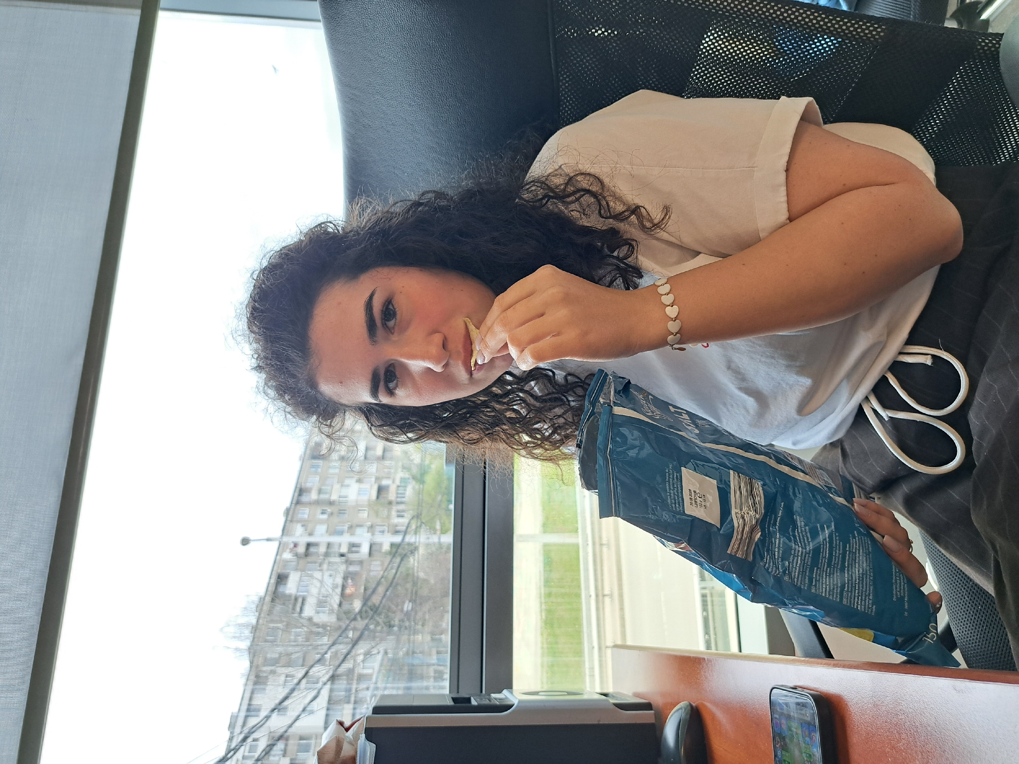 A young woman with curly dark hair is sitting in an office chair, eating chips from a blue bag. She is wearing a white t-shirt and a heart-shaped bracelet. A window is visible behind her, offering a view of a cityscape with a tall building and trees, presumably in Sofia, Bulgaria. A desk is present in front of her, with a smartphone, a black computer accessory, and what appears to be a document.