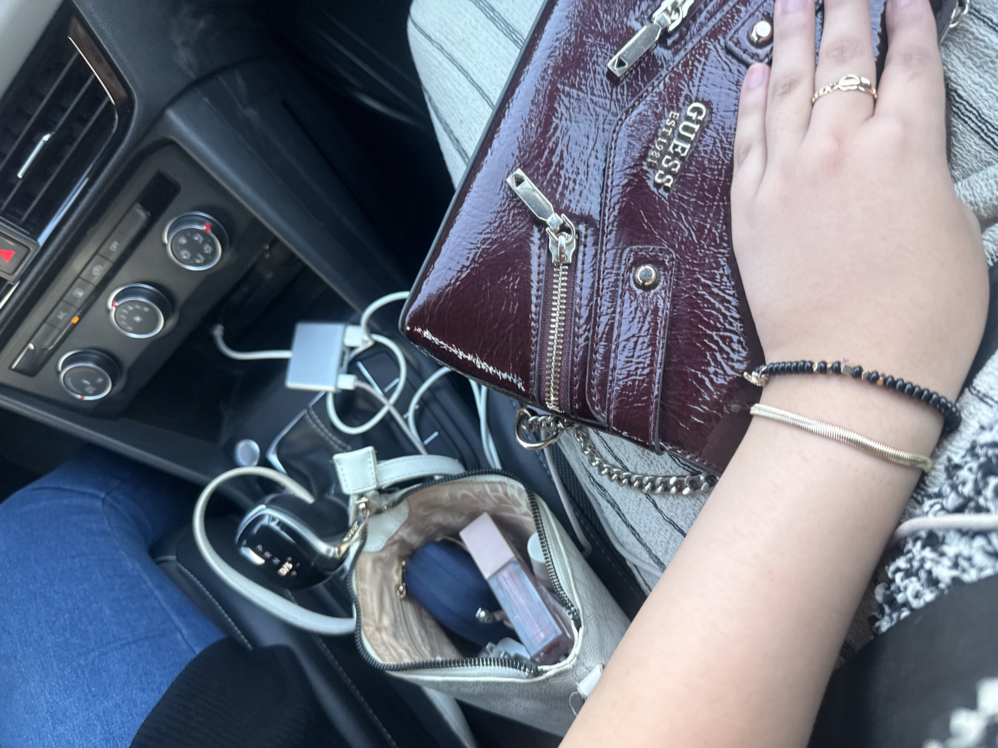 The image shows an interior car scene. On the left, part of a car's dashboard with control knobs is visible, along with the gear shift and a glimpse of the driver's leg wearing blue jeans. A white handbag containing a blue compact and a lip gloss is placed near the gear shift. The right side features a woman's hand with a gold ring and bracelets resting on a shiny, maroon Guess handbag. The woman is wearing striped pants. The location is Unknown, Unknown.