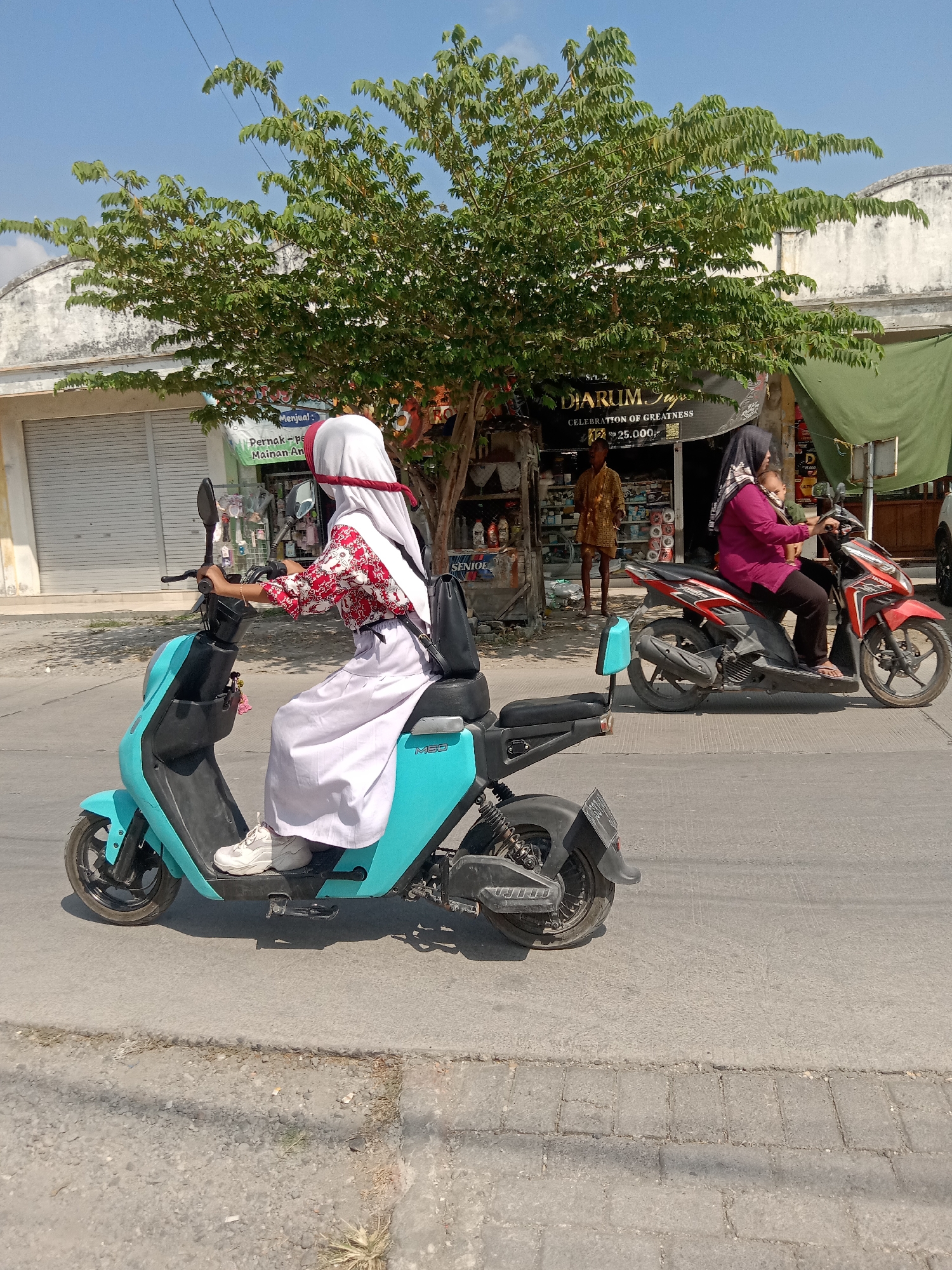 In the city of Klepek, Indonesia, a young person wearing a white hijab and a red and white patterned shirt rides a teal electric scooter. The scooter has a black seat and accents. The rider's legs are clad in a white skirt, and they are wearing white sneakers. A black backpack is slung over the rider's shoulder. Behind them, on the right side of the image, another person wearing a dark hijab and a purple top rides a red and black motorcycle. They are carrying a baby on their lap. In the background, there is a shop with a green awning and various items displayed on shelves, including what appear to be bottled products and a sign that reads "DIARUM SUGAR." A man in a yellow patterned garment stands outside the shop. The scene takes place on a paved road under a clear blue sky, with a tree providing shade in the middle ground. The buildings in the background have white walls and shuttered windows.