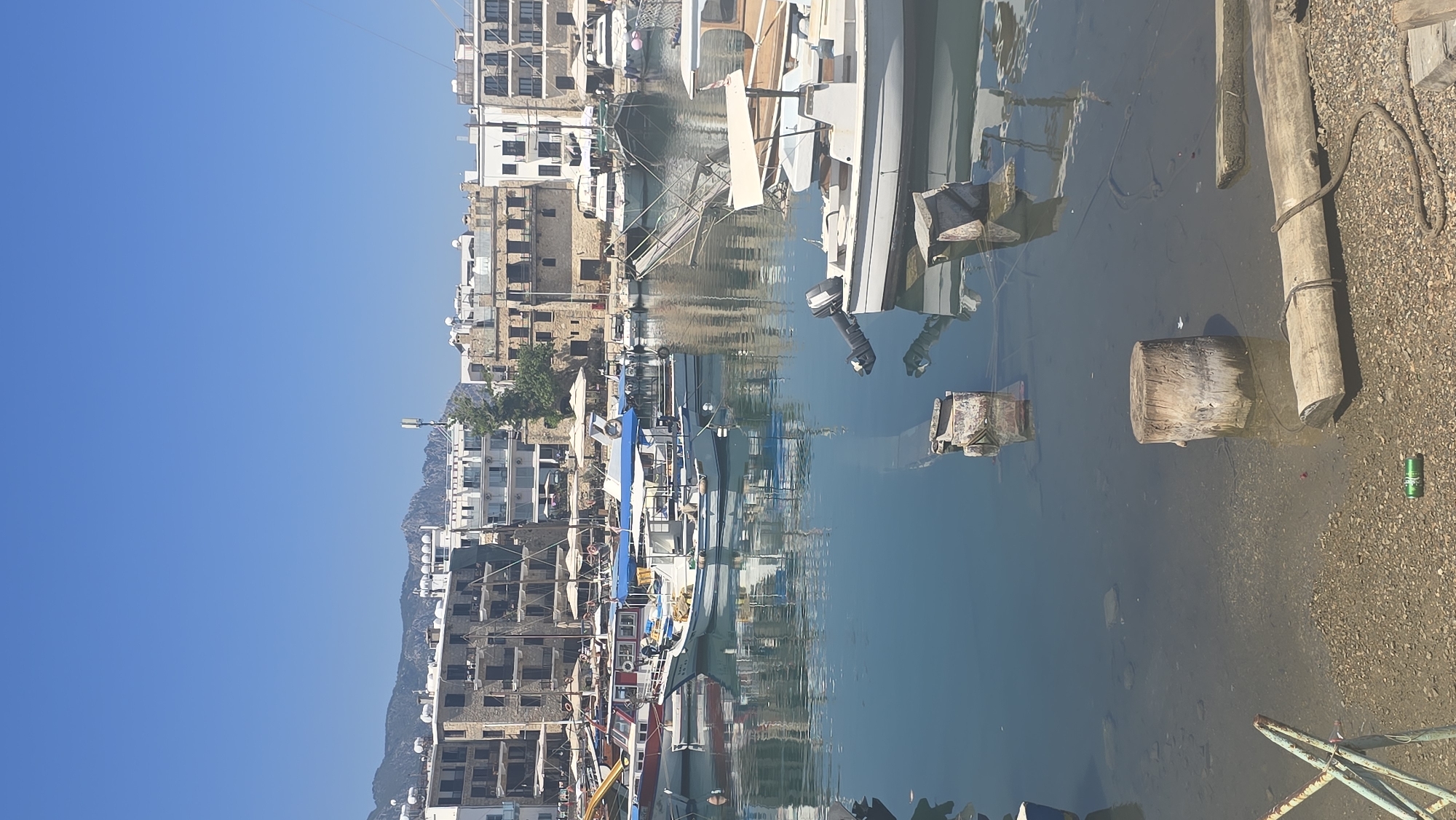 The image depicts a harbor scene in Kyrenia, Northern Cyprus. Numerous boats of various sizes are moored in the calm, blue water. Many of the boats are white with blue accents. In the background, there are several multi-story buildings with stone and white facades, characteristic of Mediterranean architecture. Some buildings have balconies and awnings. Mountains rise in the distance behind the buildings, under a clear blue sky. The foreground shows a rocky shore with a wooden log and some metal structures. The water reflects the sky and the boats.