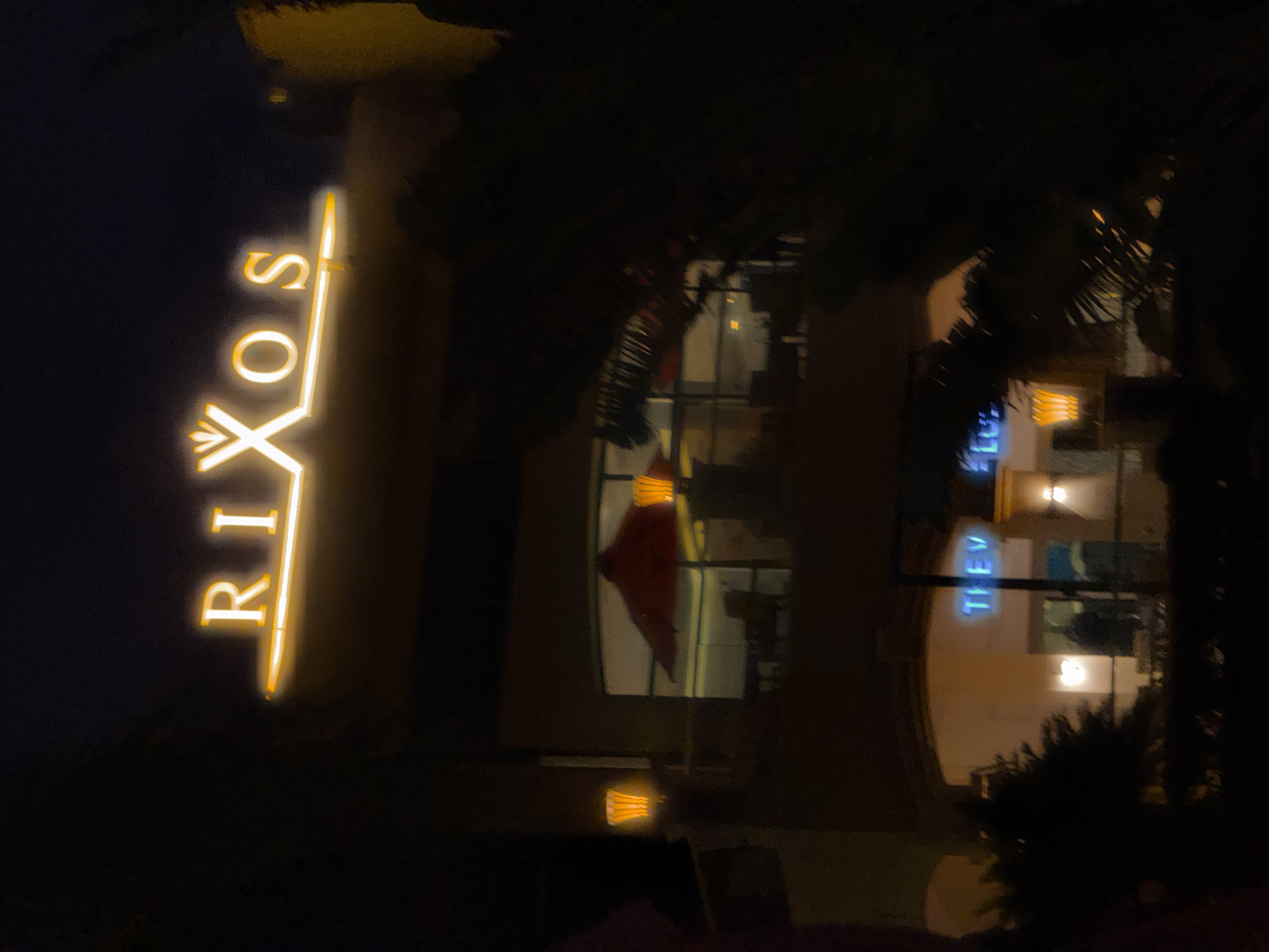 A nighttime, eye-level, medium shot shows a dimly lit building with a prominent neon sign reading "RIXOS". The sign is illuminated in a warm yellow hue and features an ornate crown-like symbol above the "X". Below the "RIXOS" sign, a balcony area is visible with outdoor seating, a red umbrella, and decorative lighting. Further down, another section of the building displays neon blue text, partially visible, reading "THE VE LU". The scene is partially obscured by dark foliage in the foreground, suggesting the photograph was taken from a distance through trees. The overall atmosphere is dark and slightly blurry, typical of a low-light photograph.