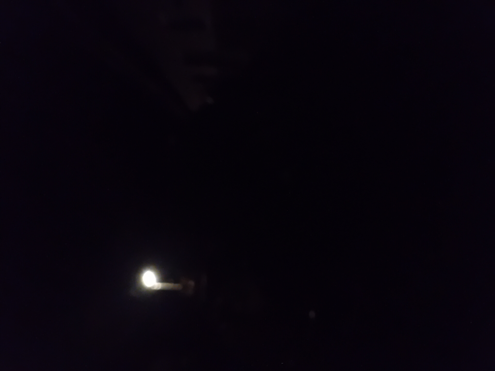 The image is a dark, nighttime photograph with a single street light illuminated in the upper left quadrant. The light source appears to be a traditional street lamp with a visible pole. The surrounding environment is obscured by darkness, with no discernible structures or significant features visible in the immediate vicinity of the street light. There is a faint suggestion of a building or structure in the background, to the right of the street light, but it is too indistinct to identify. The image appears to be taken from ground level, looking upwards towards the street light. No people or animals are visible.