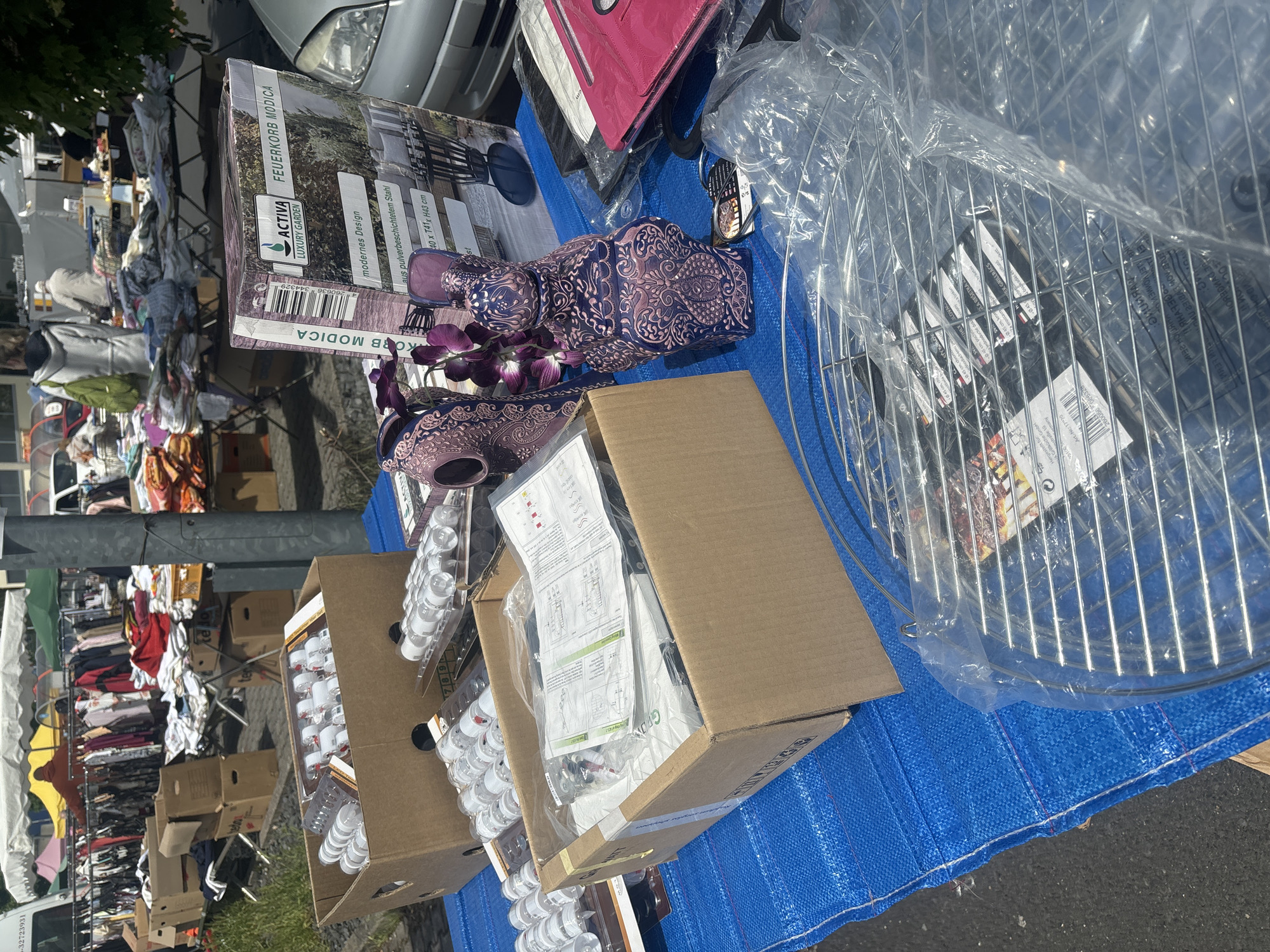 The image shows a variety of items laid out on a blue tarp, likely for sale at a flea market or outdoor bazaar.
Objects present: Boxes filled with small, white-topped containers, a box with instructions and hardware, two ornate purple vases with faux flowers, a boxed fire pit, a pink bag, and a metal grill grate still in its plastic wrapping.
Environment: The scene takes place outdoors in Eichenzell, Germany, with a glimpse of parked vehicles and a clothing rack in the background suggesting a market or sale environment.