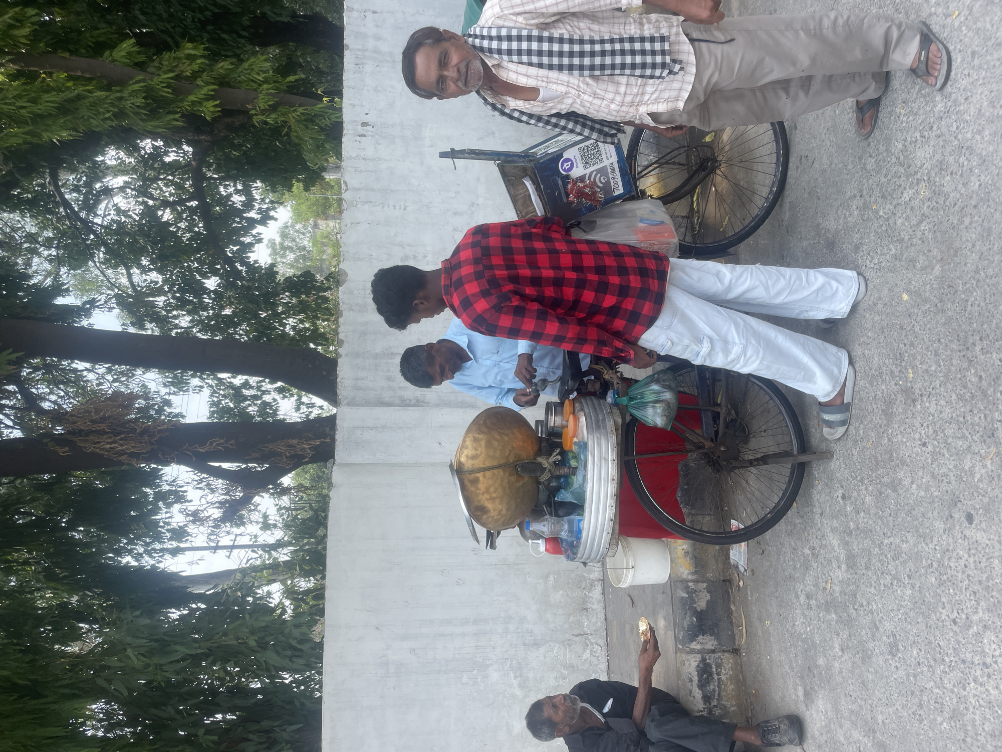 The image depicts a street scene in Pathankot, India, featuring a mobile food cart and several individuals. A vendor, wearing a blue shirt, is seen preparing food at the cart, which is mounted on a bicycle. A young man in a red and black checkered shirt and white pants stands nearby. To the left, an older man in a black shirt and pants is seated, holding a small plate. On the right, another man wearing a checkered scarf and light-colored pants stands and looks towards the camera. The food cart is equipped with various containers and bottles, including a large brass pot. The environment includes a white wall in the background, foliage from trees overhead, and a paved street. The overall impression is of a typical street food vendor setup in an urban Indian setting.