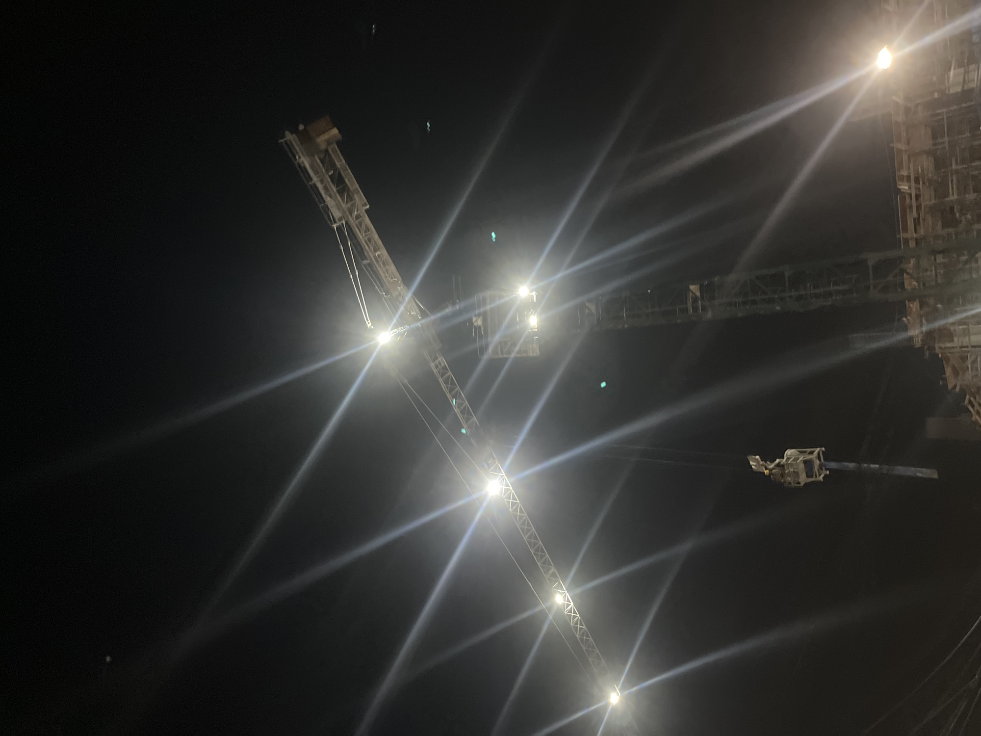 An outdoor night-time, low-angle, close-up shot of a construction crane is captured against a dark sky. The crane, constructed of metal, extends diagonally from the lower left to the upper right of the frame, its structure illuminated by several bright lights that create prominent starbursts and light flares. A smaller, enclosed cabin structure is visible near the top of the crane. Below the main crane, a section of a second, taller crane, possibly under construction, is visible, also lit by bright lights. In the lower portion of the image, the outlines of unfinished construction work with scaffolding and wooden structures are dimly discernible, also illuminated by a bright light in the lower right corner. The overall scene depicts an active construction site at night in Tangerang, Indonesia.