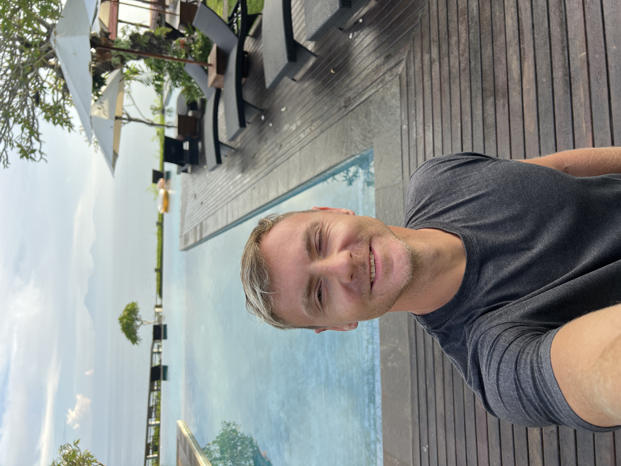 A man is captured in a selfie, positioned on the left side of the frame, smiling at the camera. He has light skin, short blonde hair with darker roots, and light stubble, and is wearing a dark grey t-shirt. The man appears relaxed and happy.
Behind and to his right is a large swimming pool with light blue water, possibly an infinity pool, which extends into the distant background. The pool is bordered by dark wooden decking, visible in the foreground and continuing around the pool area. On the decking around the pool, several dark lounge chairs and white sun umbrellas are arranged. Beyond the pool, the scene opens to a scenic view under a bright, mostly clear sky with subtle clouds. Lush green trees and foliage are visible both in the near and far background, with one prominent small tree reflected in the pool water in the distance. A faint, small orange object can be seen floating in the pool in the background. The overall setting suggests a tropical resort or hotel environment, aligning with the provided location context of Kuta Selatan, Indonesia. The lighting indicates it is daytime with good weather conditions. No text is visible in the image.