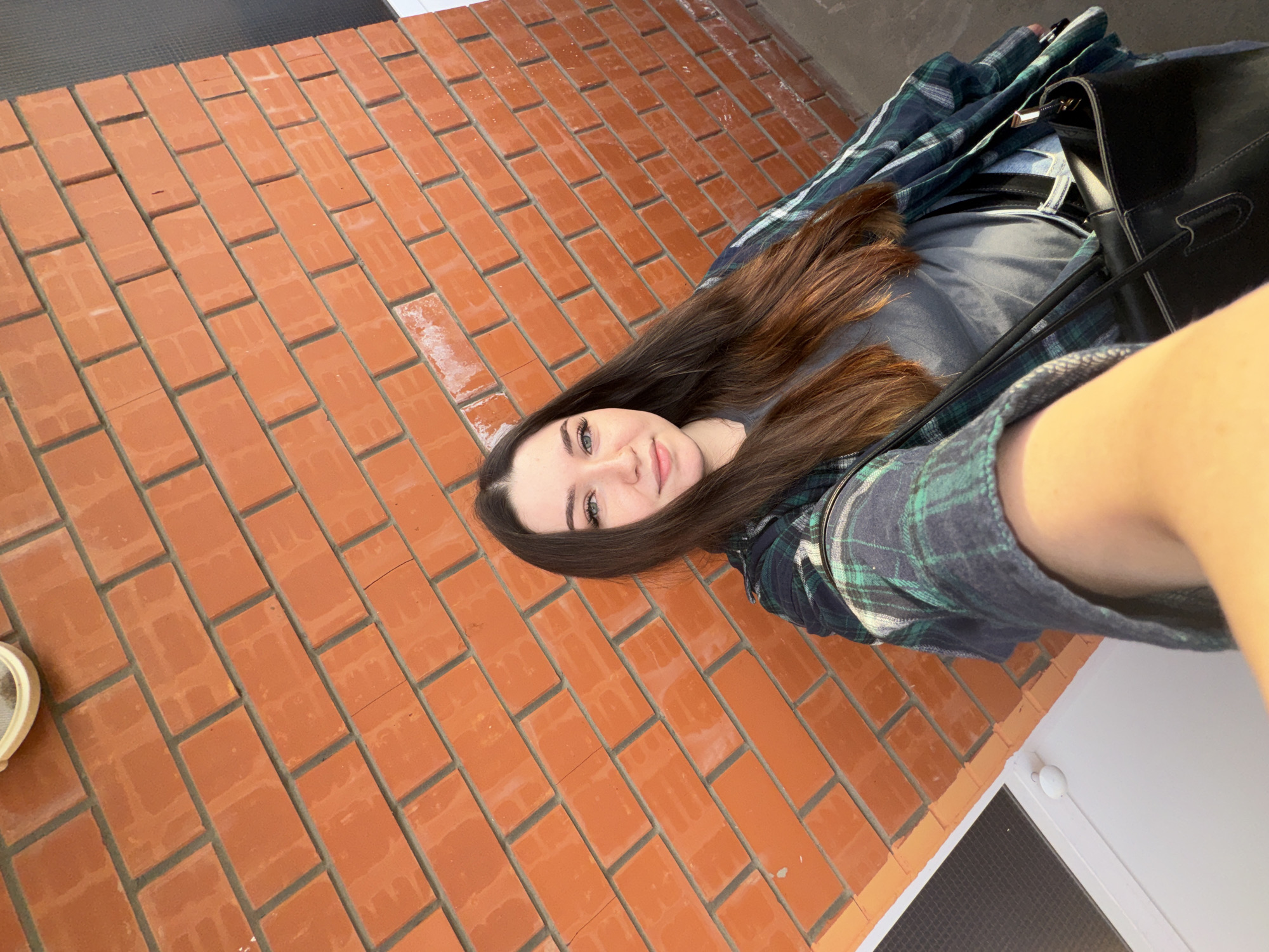 A young woman with long, dark brown hair and striking blue eyes takes a selfie against an orange brick wall. She is wearing a grey t-shirt and an open, plaid flannel shirt in shades of blue and green. A black handbag with a gold zipper hangs from her shoulder. The background is a textured orange brick wall with visible mortar lines. The lighting suggests an outdoor setting, possibly during the day, with some sunlight casting shadows on the wall. To the left of the woman, a white door frame and a small white light fixture are partially visible. The overall scene is casual and urban.